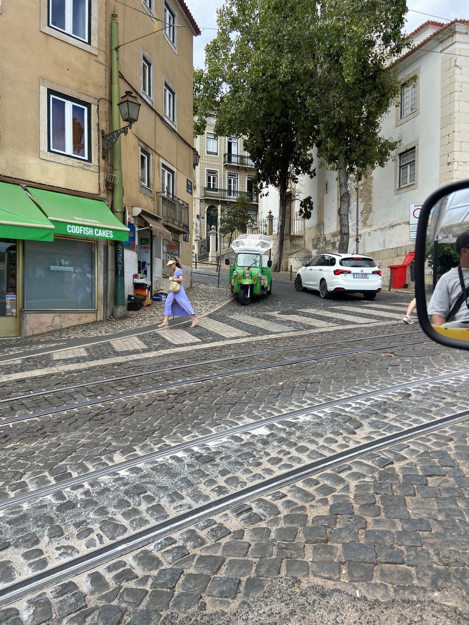 A street scene showing cobblestone pavement with tram tracks, a woman in a light purple dress walking, a small green motorized vehicle, cars, trees, buildings with balconies, and a vendor stall with a green awning that reads 'CODFISH CAKES.' A half-visible man is in the side rearview mirror.