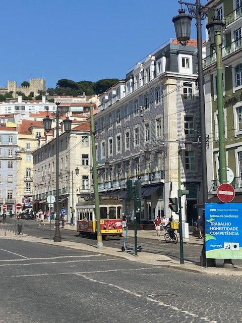 Street scene in Lisbon, Portugal, with a tram, pedestrians, colorful buildings, and a clear blue sky.