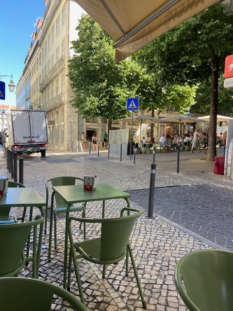 Outdoor cafe seating area with green chairs and tables, under umbrellas, on a cobblestone street in a city with trees and buildings.