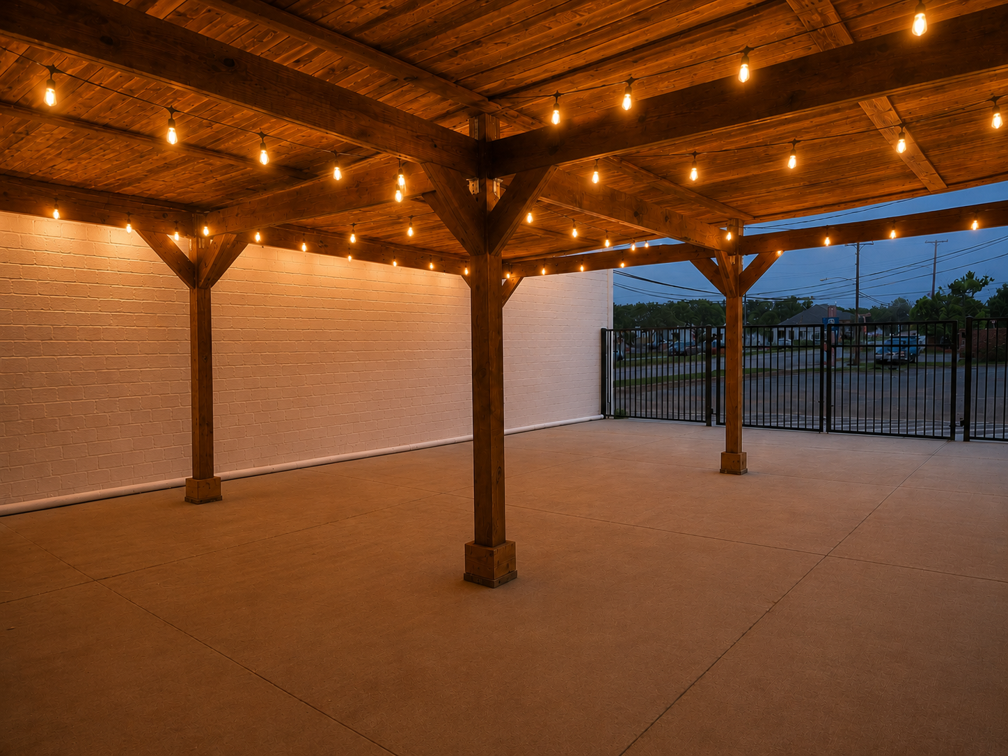 An outdoor patio area with a wooden pergola decorated with string lights, a white brick wall, and a gated boundary overlooking a parking lot at dusk.
