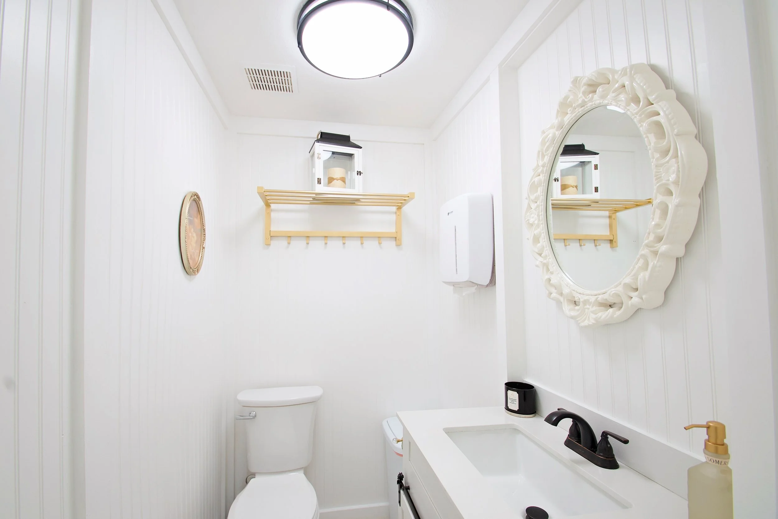 A small bathroom with white paneling walls, a white toilet, a white sink with a black faucet, a black trash can, a white wall-mounted cabinet with glass doors, a wooden towel rack, a round mirror with an ornate white frame, a small framed picture, a 