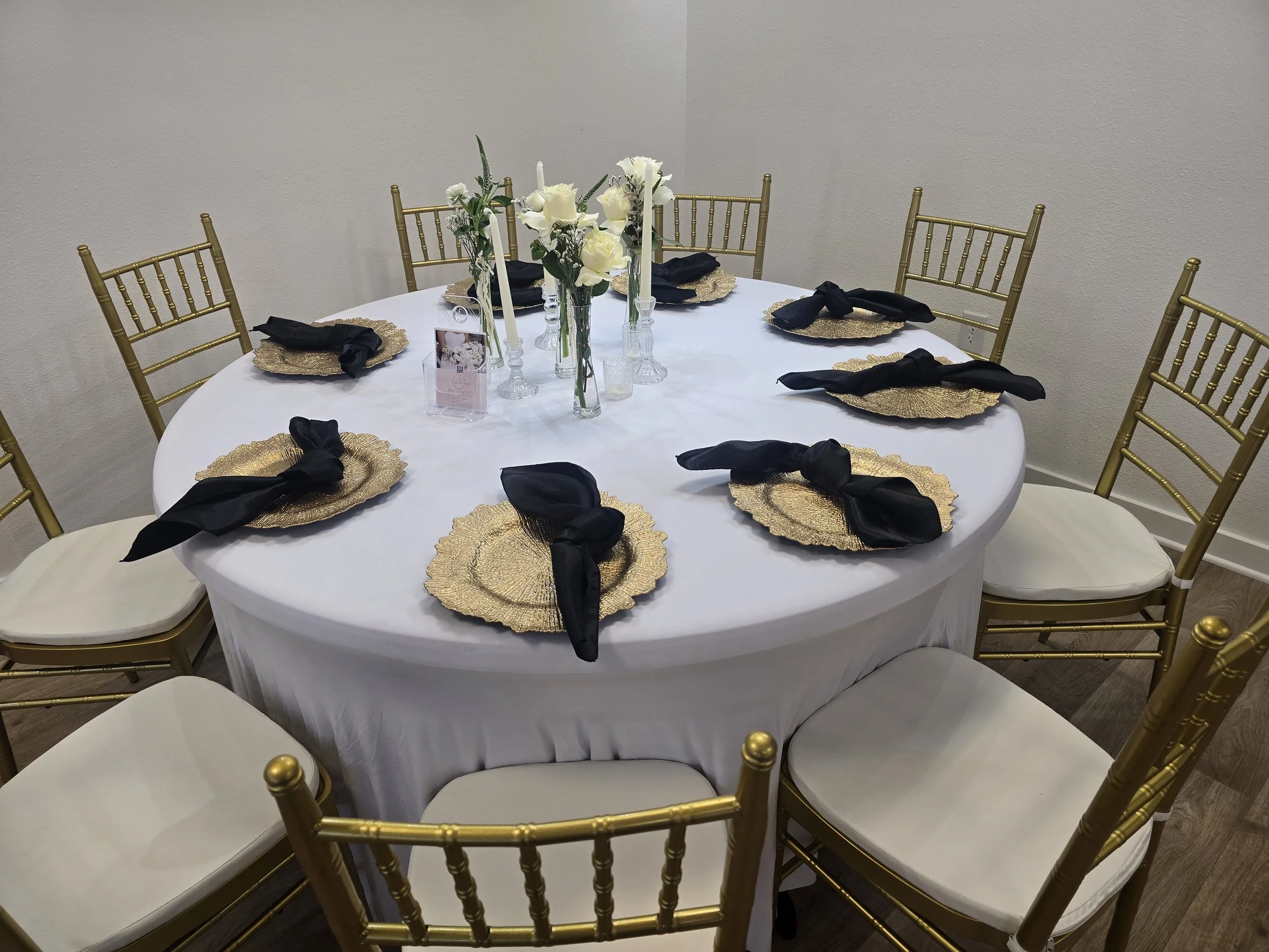 Round table with a white tablecloth, gold chairs, black napkins on gold plates, floral centerpiece with white roses and greenery, and candle holders with tall white candles.