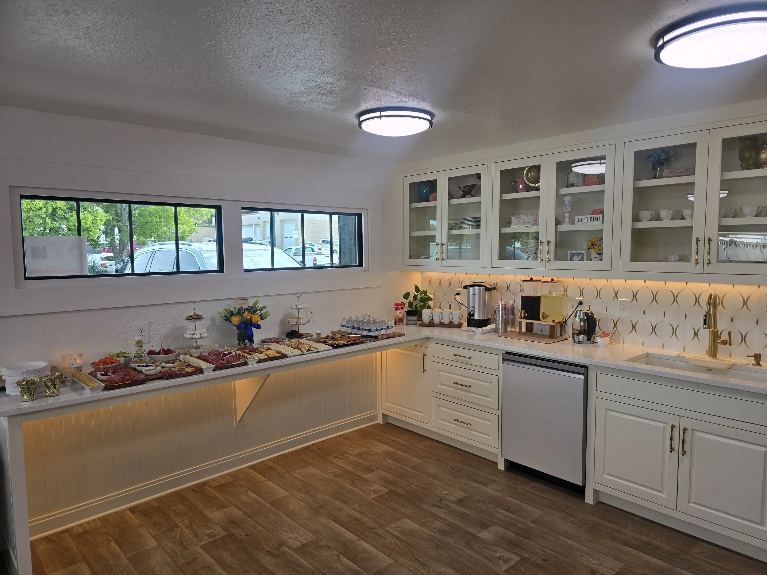 A breakfast or snack buffet setup on a white kitchen counter with various foods including fruits and bread, in a clean, well-lit kitchen with white cabinets and a large window overlooking a parking lot.
