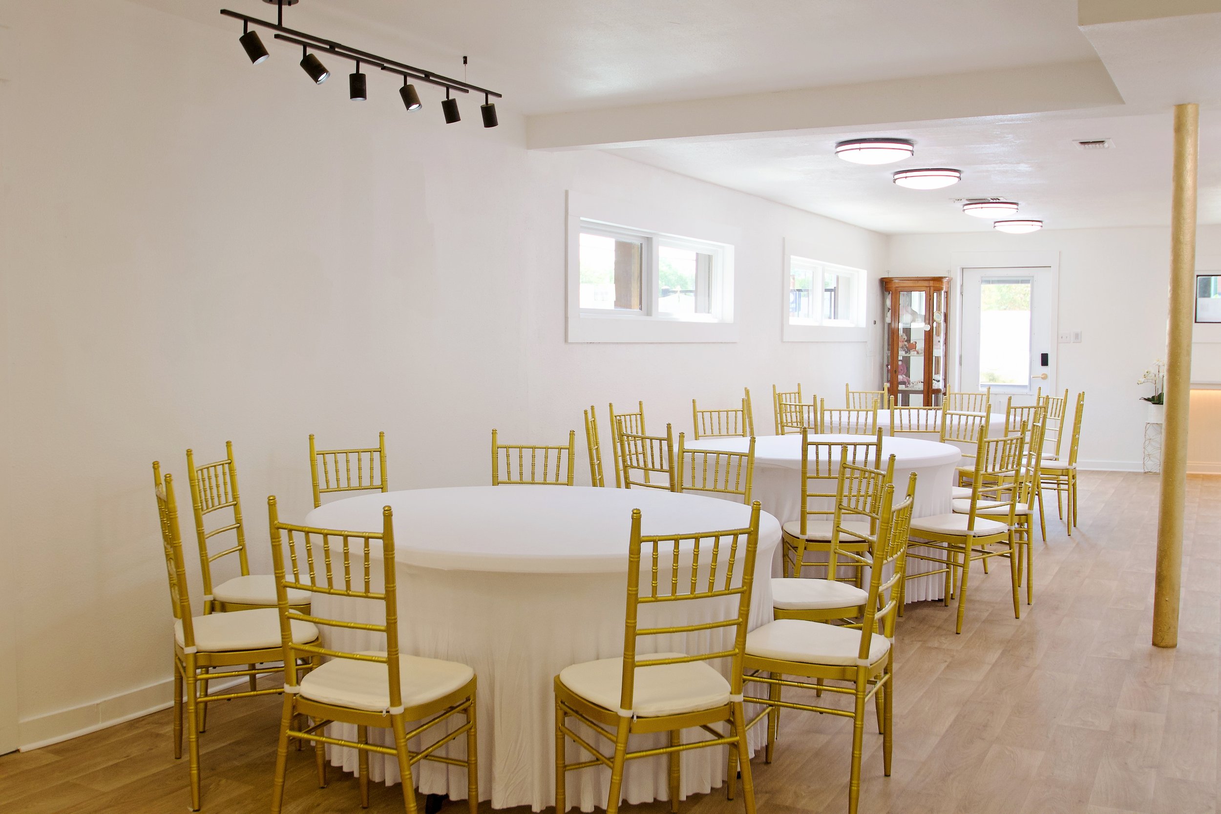 Empty banquet room with round tables covered in white tablecloths and gold chairs, large windows along the walls, and ceiling lights.