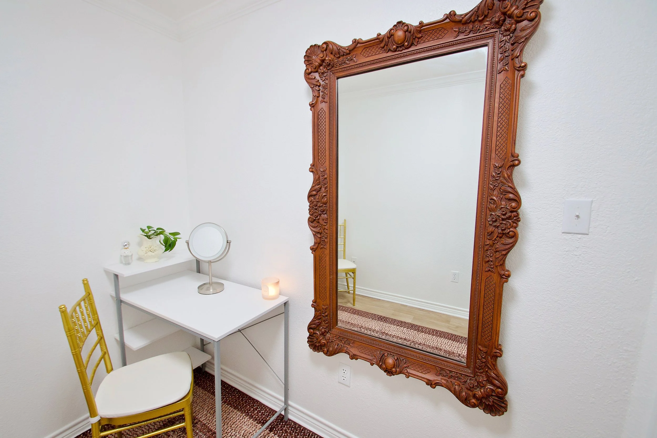 A large ornate wooden mirror hanging on a white wall, with a gold chair and a small white vanity table with a round mirror and decorative items underneath.
