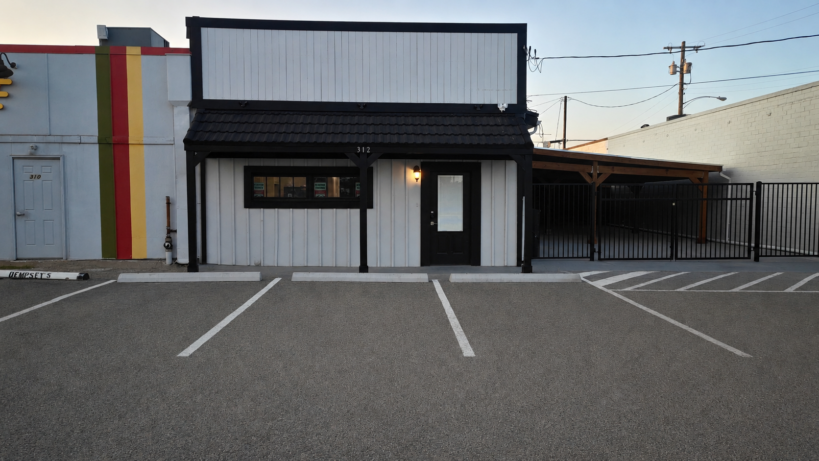 A small building with black and white exterior, a porch with black wood support beams, and a black door with a window. It has parking spaces in front, marked with white lines, and a black fence section to the right. The sky is clear and power lines are visible above.
