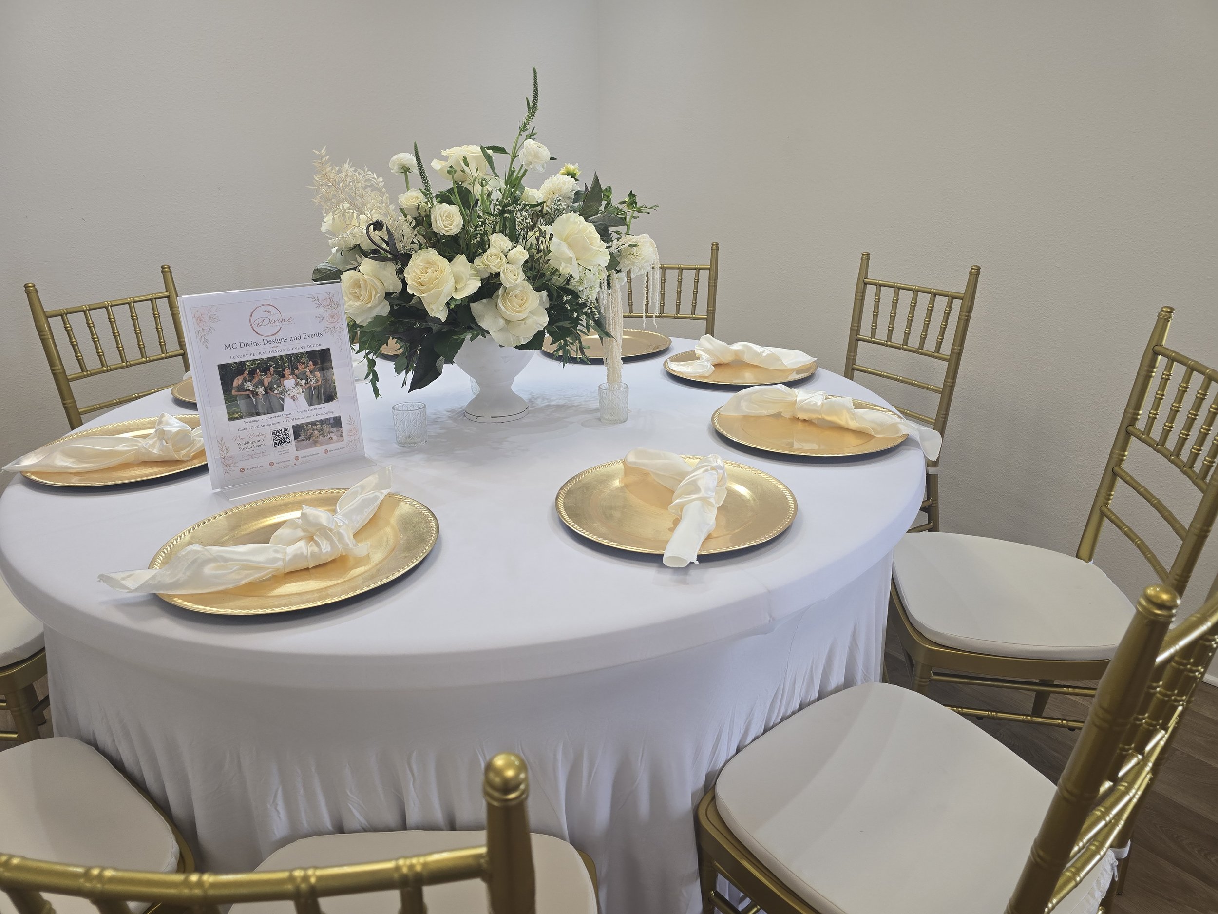 Round banquet table covered with a white tablecloth, set with gold chargers, white napkins, and decorated with a large floral arrangement of white roses and greenery. Golden chairs surround the table. A framed sign with floral design and QR code is a