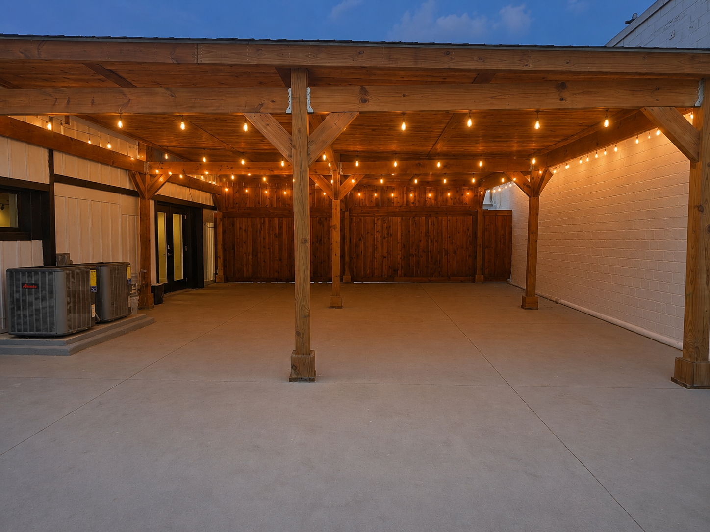 Covered outdoor patio area with string lights hanging from a wooden pergola, concrete floor, and a white brick wall on the right side.