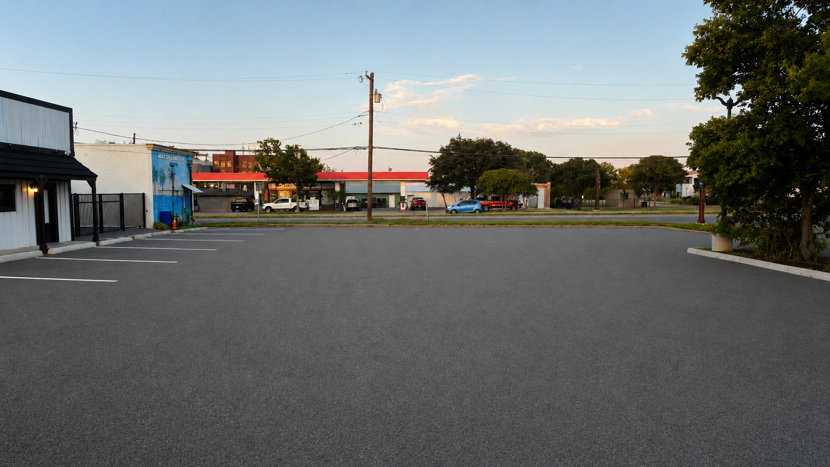 Empty parking lot with white lines marking parking spaces, adjacent to a building on the left, and trees and a street with cars in the background under a clear sky.