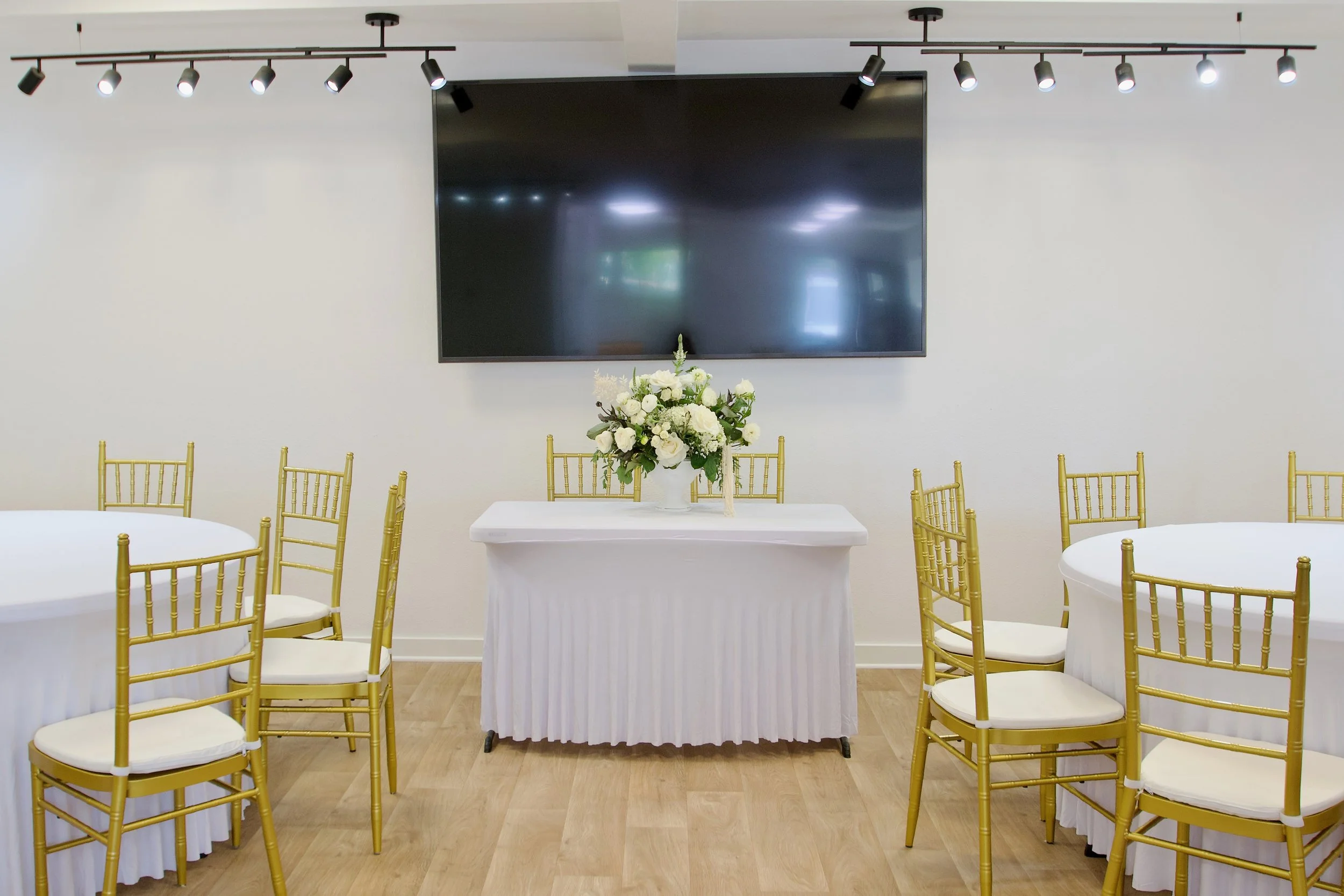 A ceremony setup with gold chairs and a floral centerpiece on a white table, against a wall with a large black screen and floral arrangement, in a well-lit room.