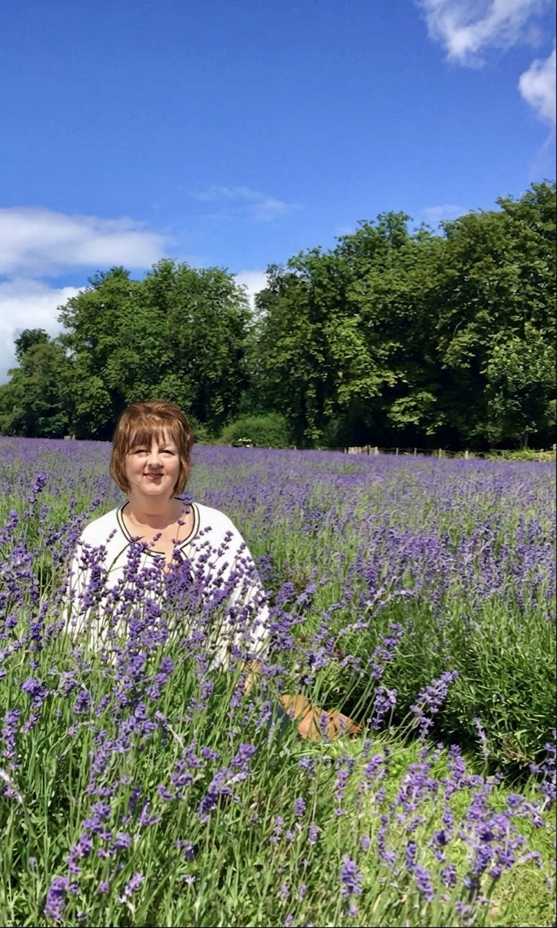 A smiling woman sitting in a field of blooming lavender flowers on a sunny day, with green trees and a blue sky with some clouds in the background.