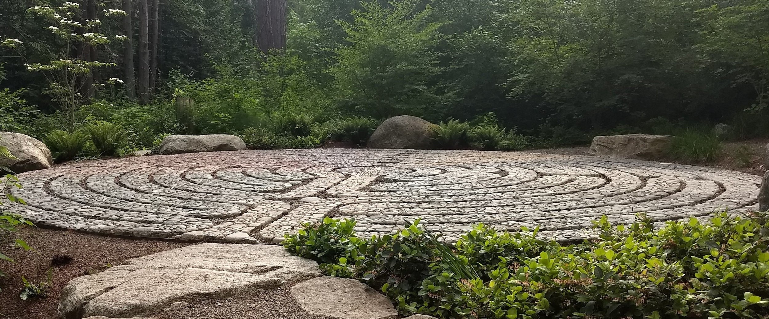 A circular stone pathway in a green outdoor setting surrounded by plants, trees, and rocks.