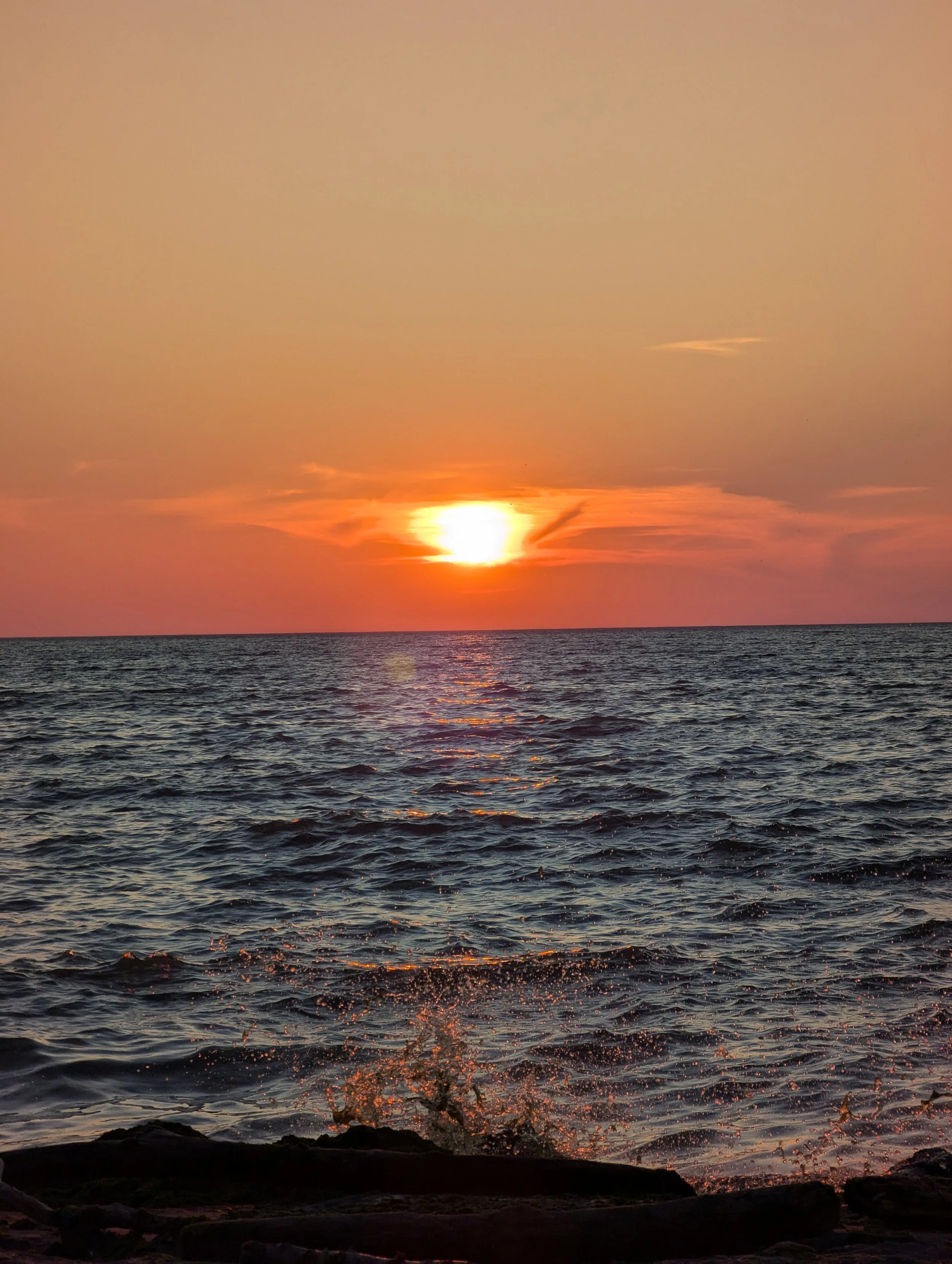 Sunset over the ocean with orange and pink sky, waves crashing near the rocky shoreline.