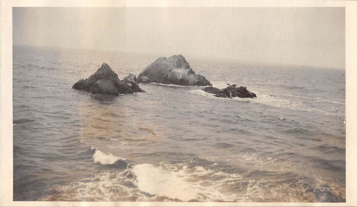 A black and white photograph of a seascape with two large rocky islands in the distance and water splashing around them.