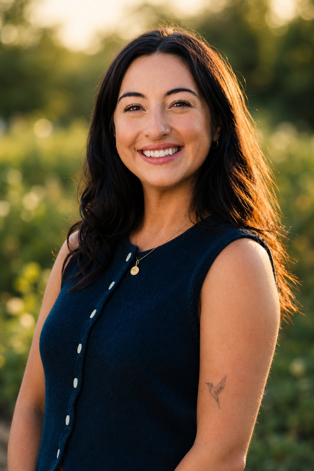 A young woman with long dark hair and a bright smile, wearing a sleeveless navy top and a gold necklace, standing outdoors during golden hour with a blurred green background.
