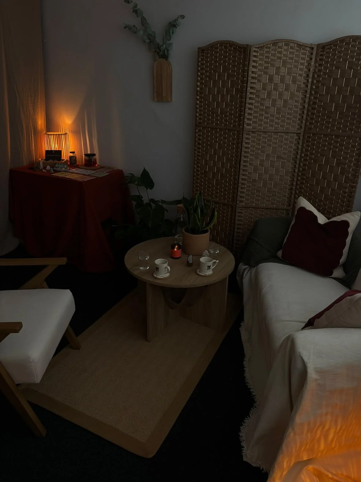 Cozy room with a white sofa, a wooden coffee table with cups and small candles, a foldable chair, a red-covered table with warm lighting, plants, and a woven room divider.