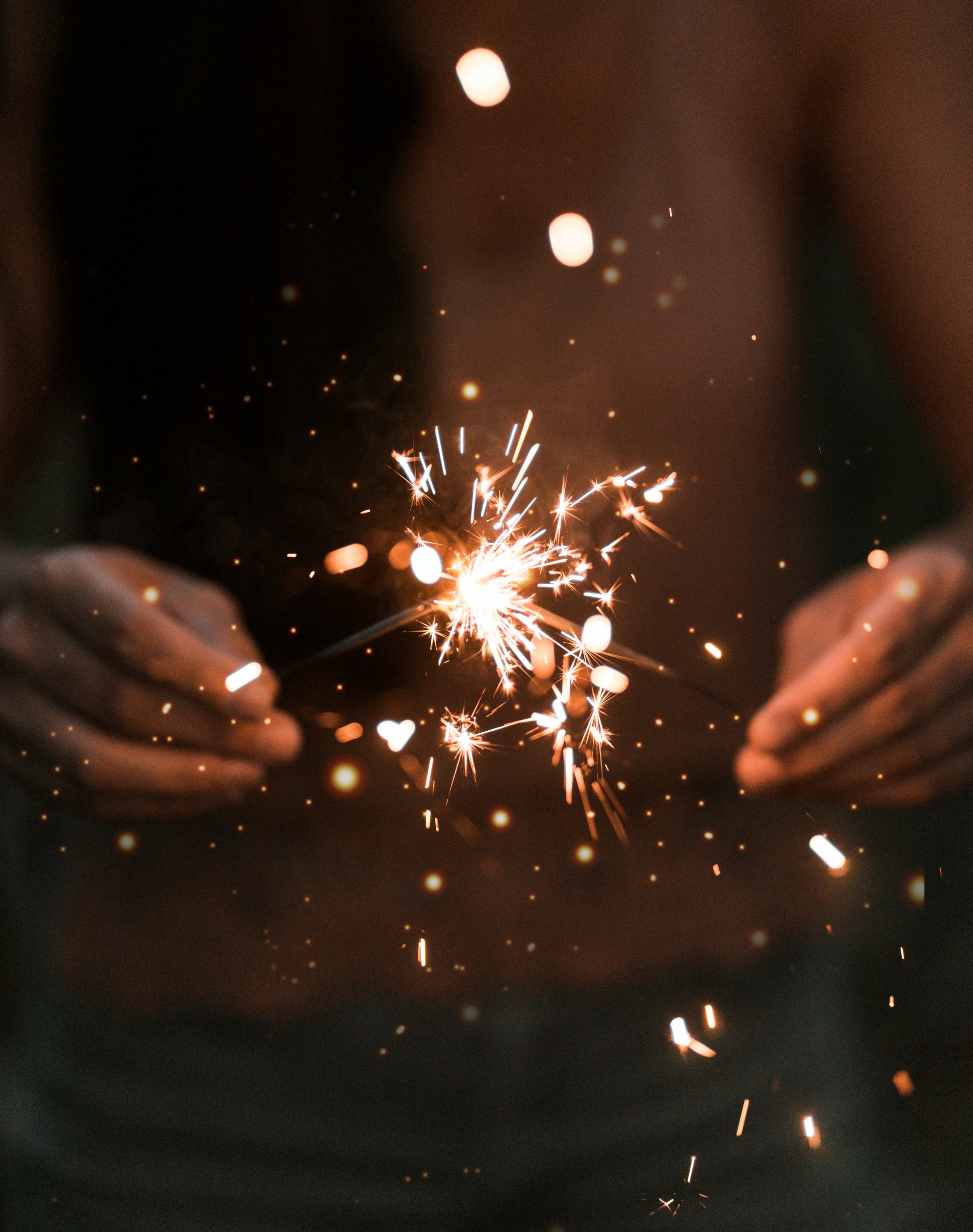 Hands holding a sparkler emitting bright sparks against a dark background.