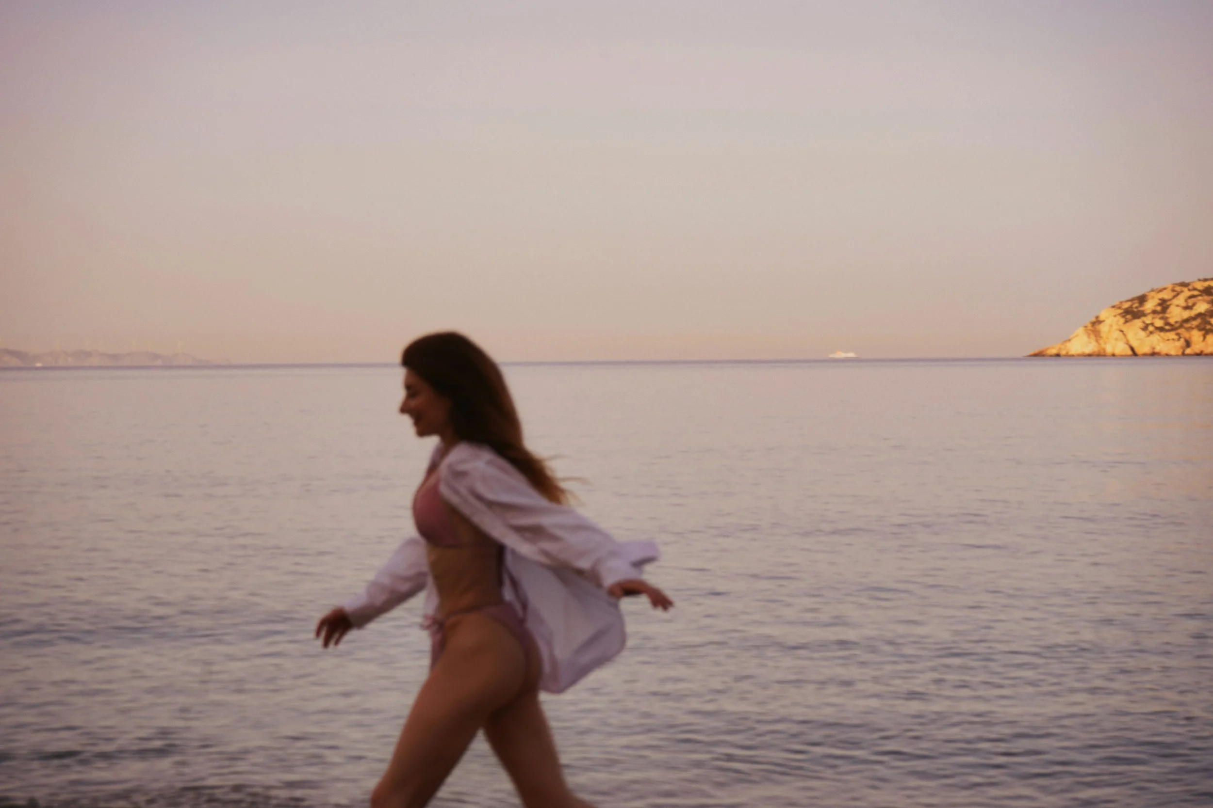 A woman running on a beach during sunset or sunrise with water and a rocky landform in the background.