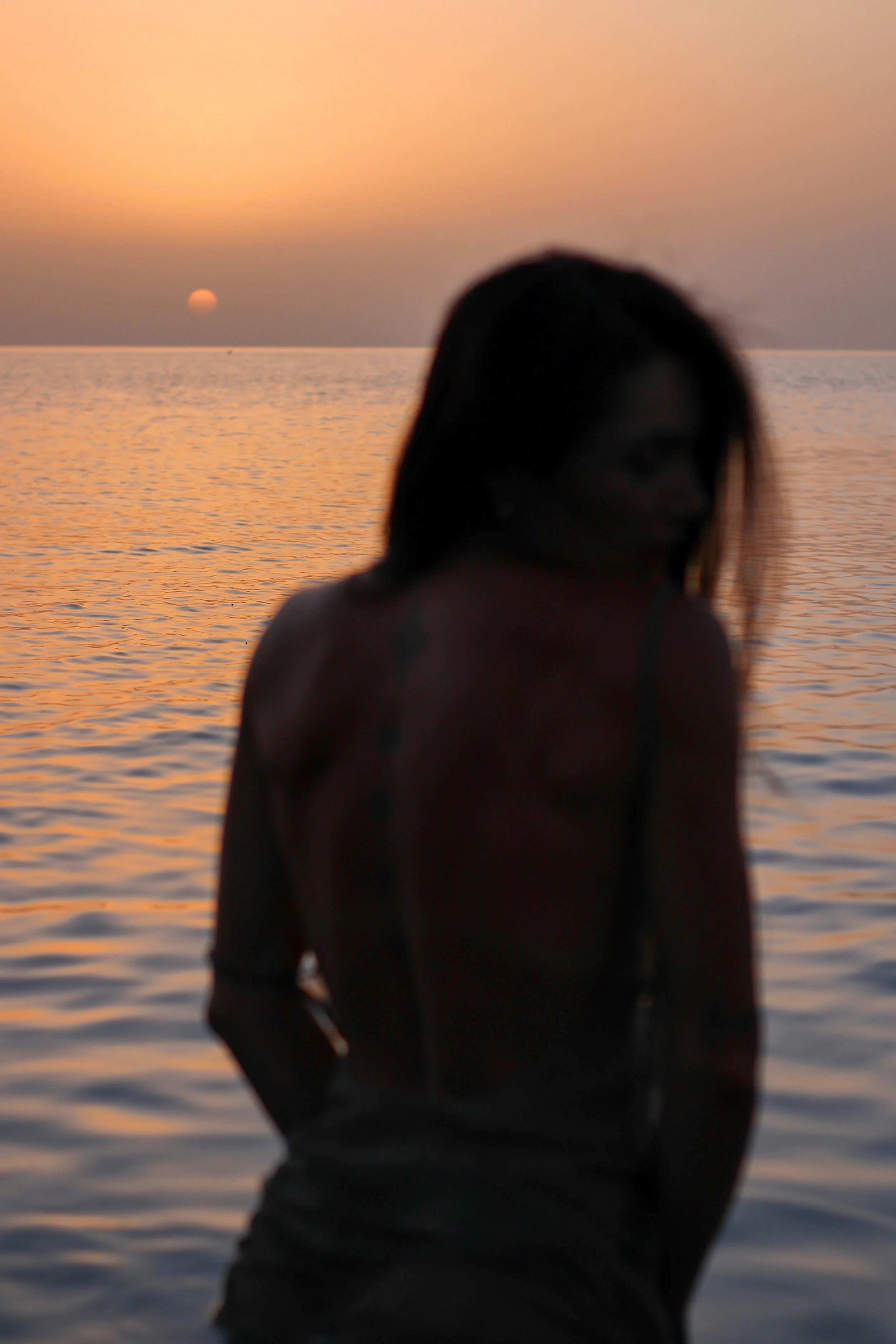 A person with long hair standing by the water during sunset, with the sun near the horizon and the sky painted with soft pastel colors.