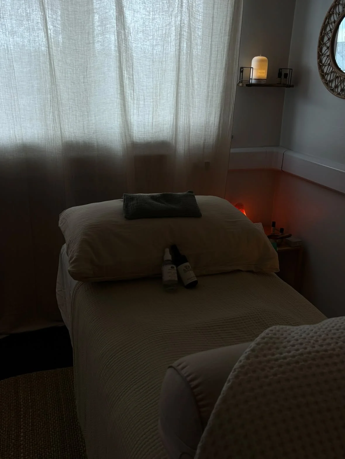 A dimly lit massage room with a massage table covered with a beige cloth. Two bottles and a towel are placed on the pillow, and a gray towel is folded on top. There are white curtains on a window in the background, with a small shelf containing a lamp and some electrical outlets, and a round mirror on the wall.