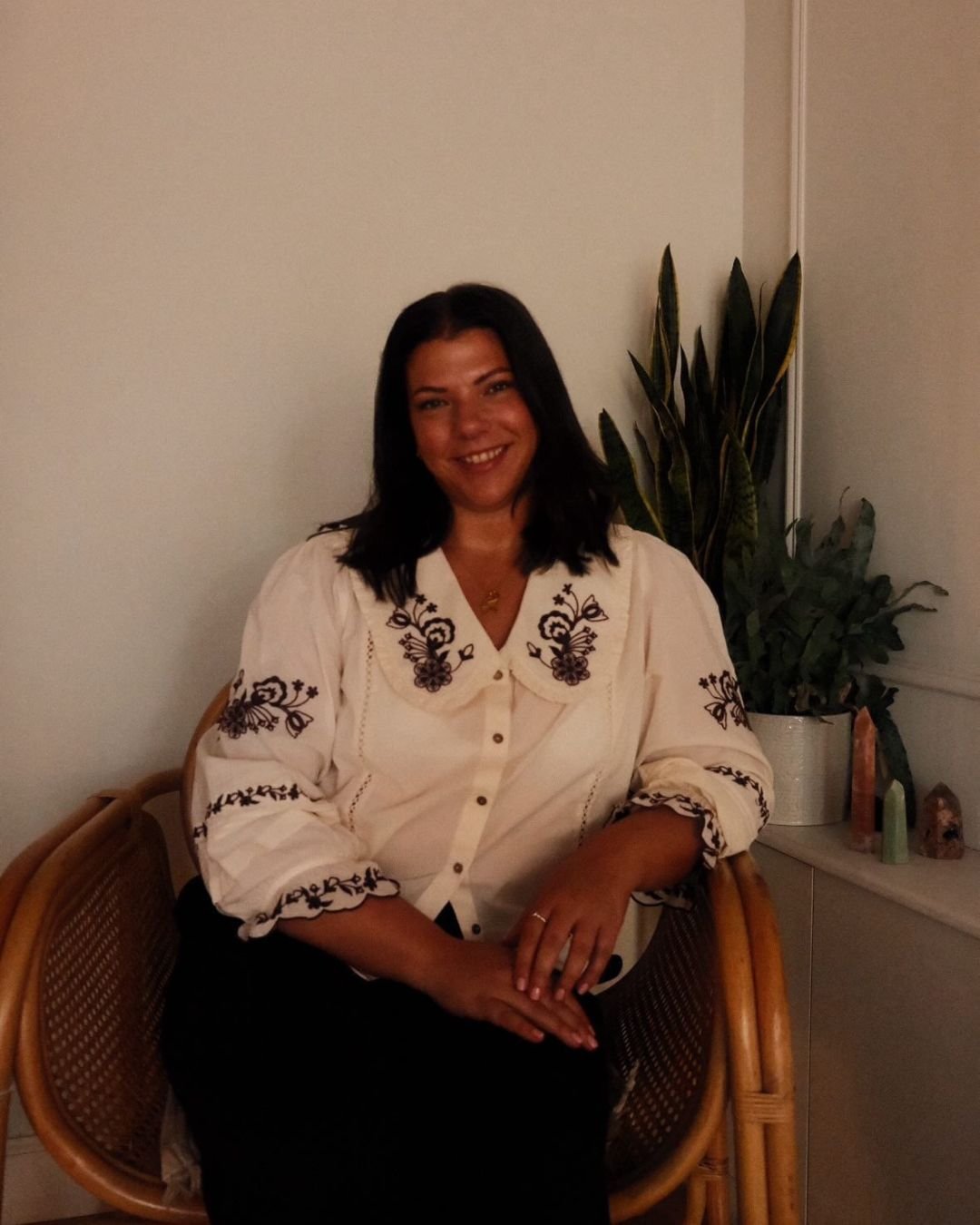 A woman with black hair and a light-colored embroidered blouse sitting on a wicker chair in a room with a potted plant and decorative items in the background.