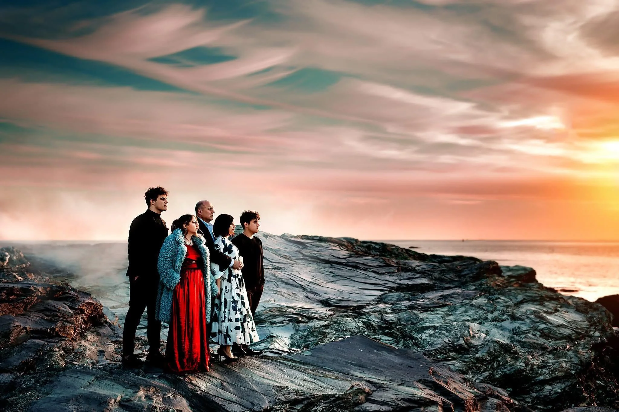 A group of seven people standing on rocks by the ocean at sunset, with layered, colorful clouds in the sky.
