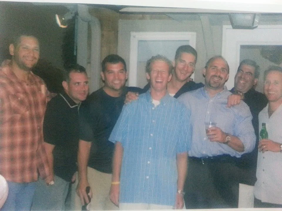 Group of eight men smiling, standing close together, some holding drinks, in a cozy indoor setting.