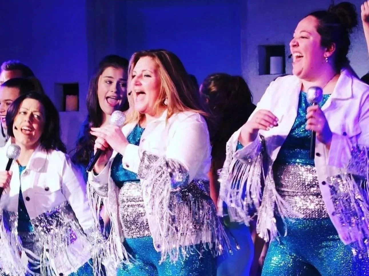 Group of women singing and dancing in sparkly blue outfits with fringe, holding microphones, enjoying a performance or karaoke night on stage.