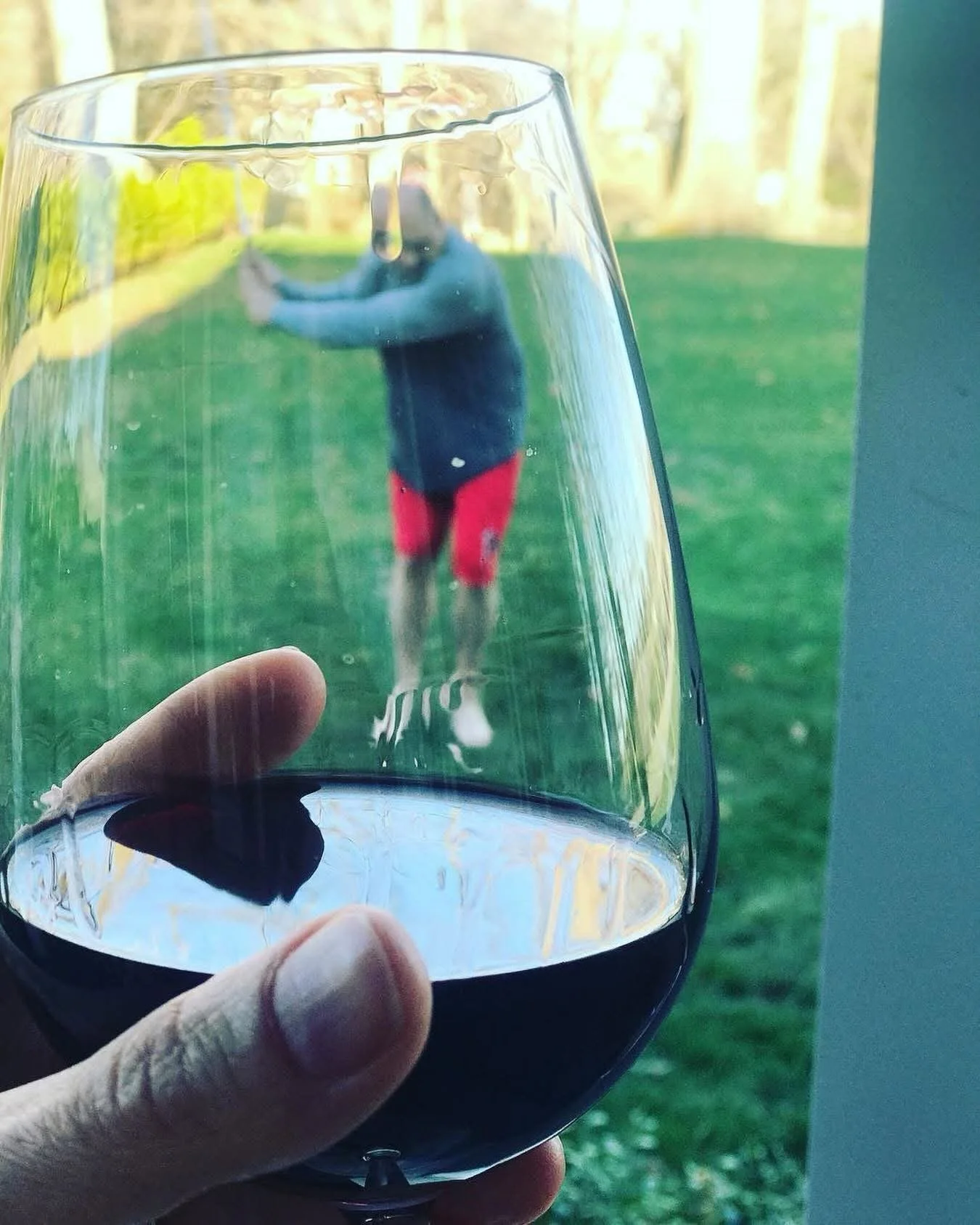 A hand holding a glass of red wine with a photograph of a person wearing red shorts and a blue shirt, leaning against a fence, visible through the glass.
