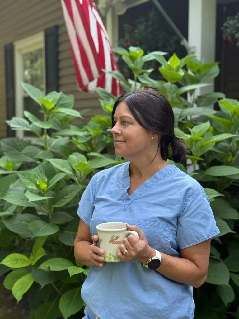A woman in blue scrubs holding a mug with a bunny design, standing in front of lush green plants with an American flag in the background.