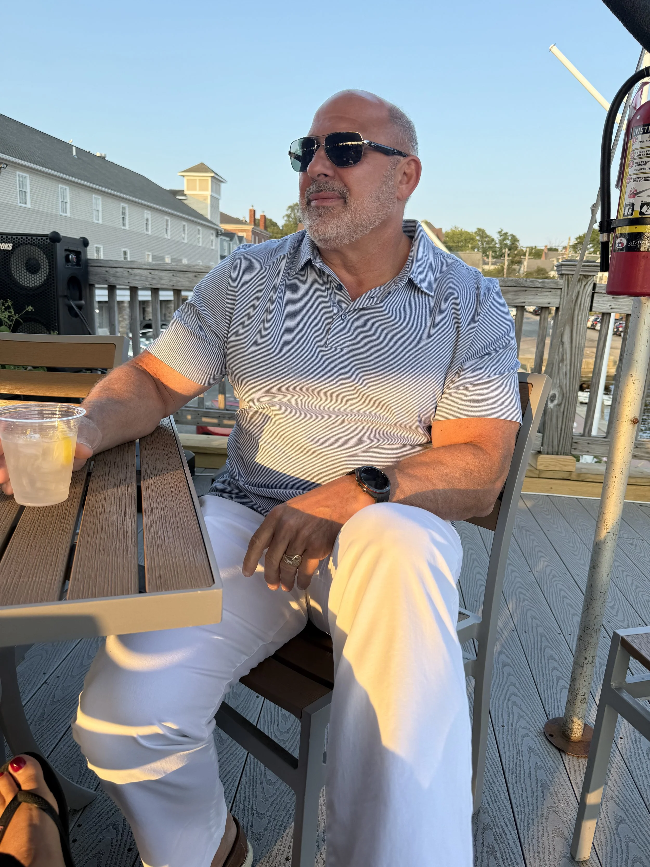 A man with sunglasses and a gray beard sitting at an outdoor wooden table on a deck, holding a clear cup with a yellow beverage, with buildings and trees in the background during daytime.
