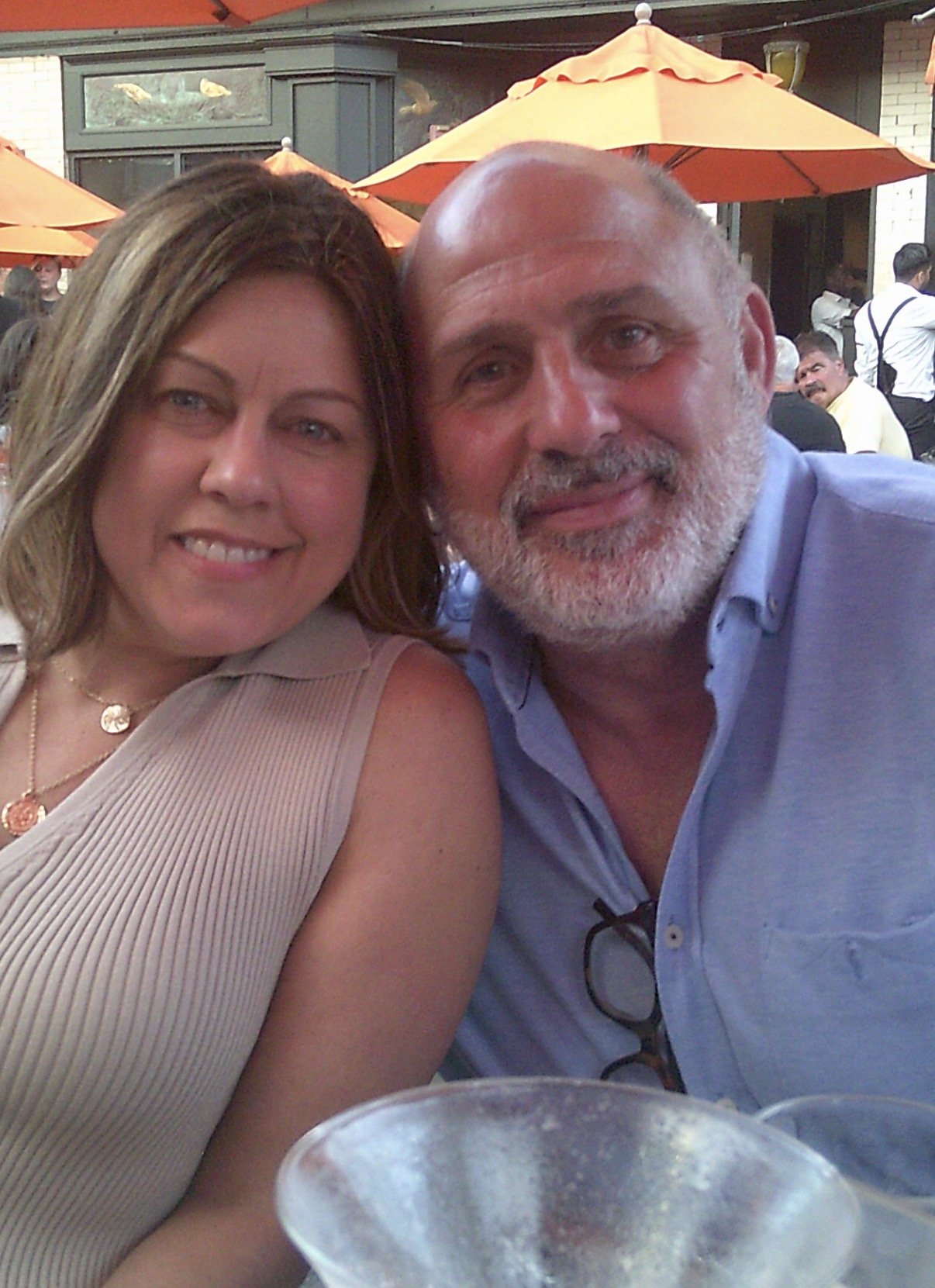 A woman and a man sitting closely together at an outdoor restaurant or bar, with orange umbrellas and other people in the background.