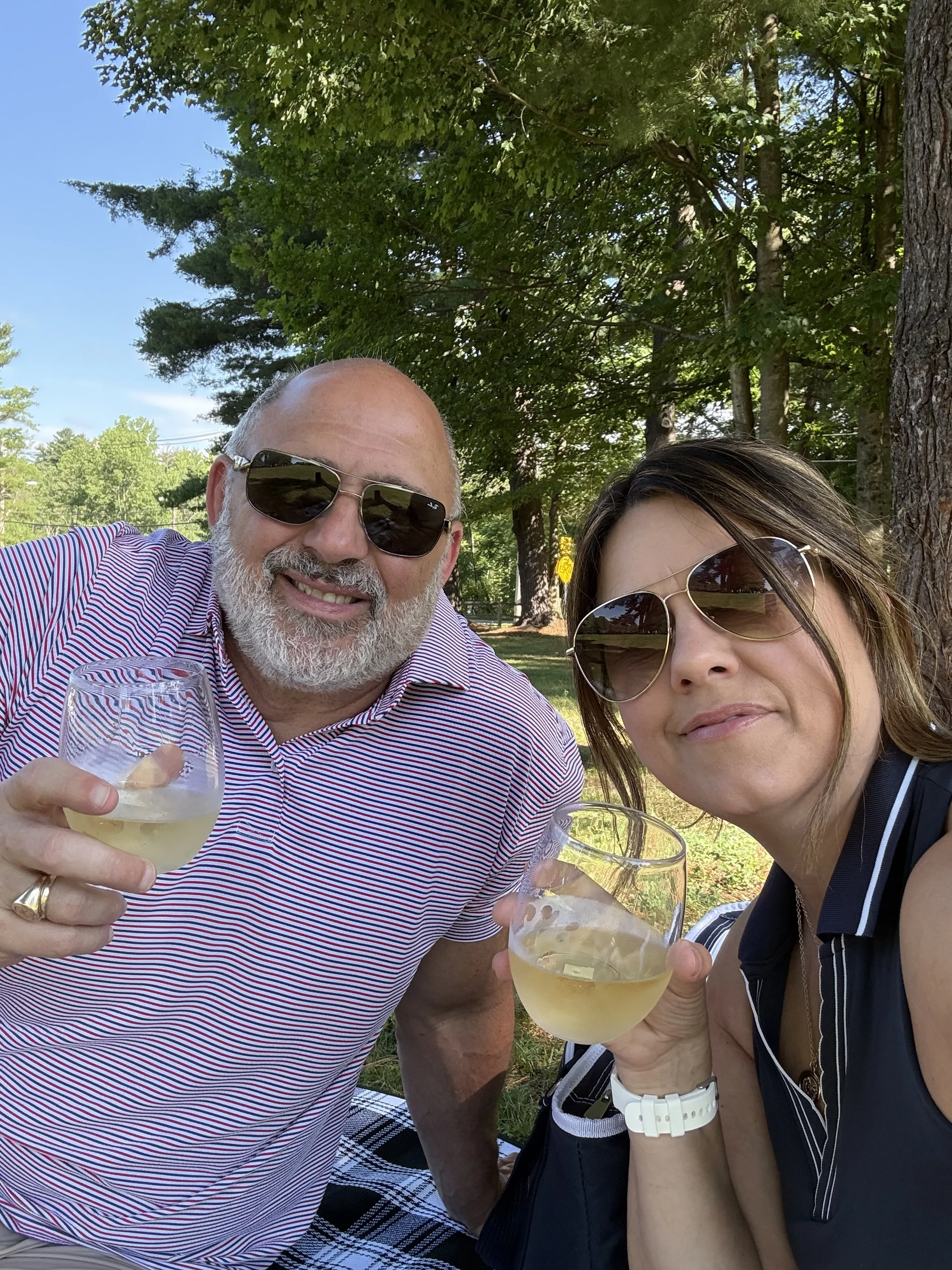 Two people outdoors holding glasses of white wine, smiling, with trees and a blue sky in the background.