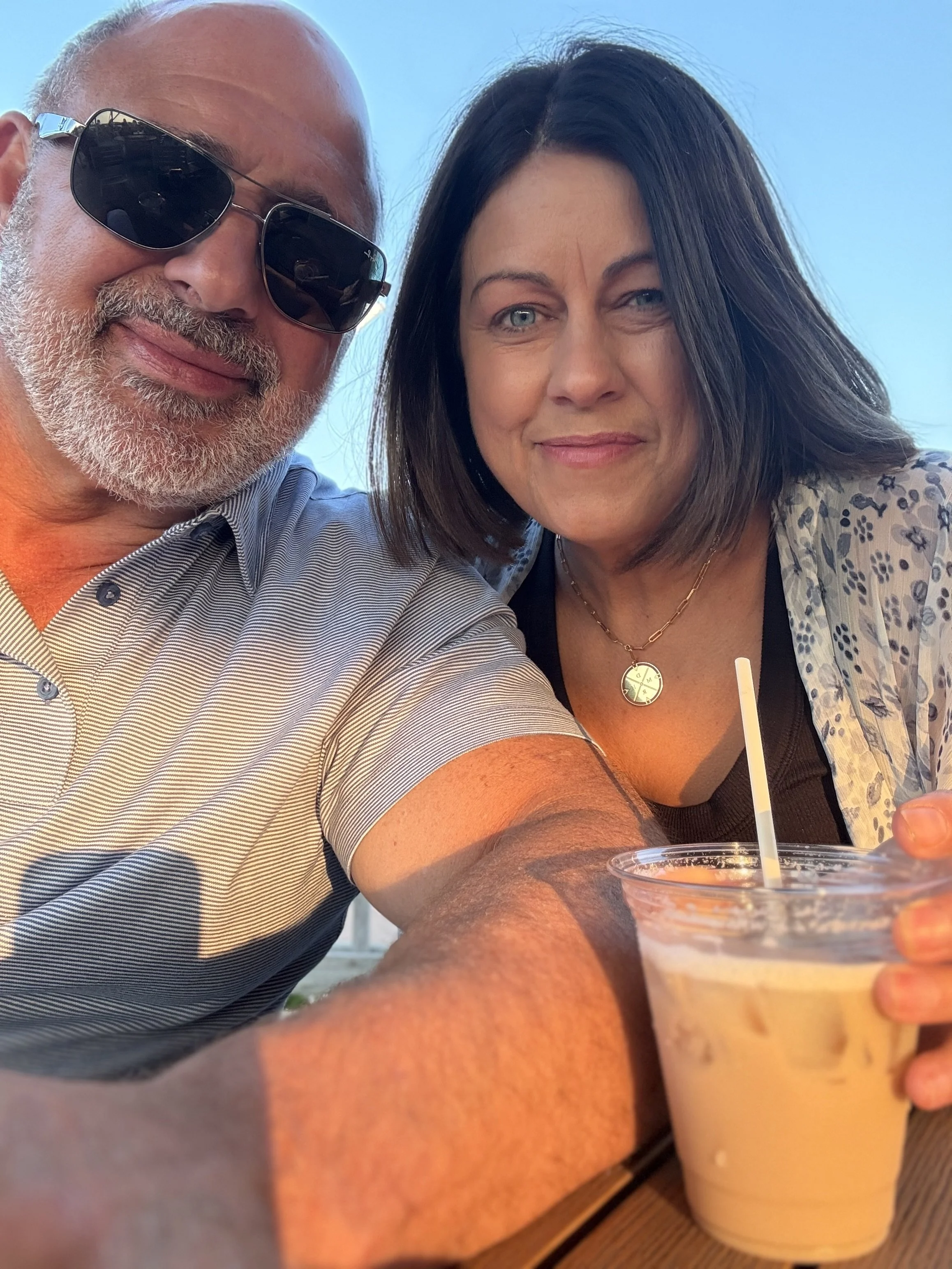 A man wearing sunglasses and a striped shirt taking a selfie with a woman with dark hair and blue eyes at an outdoor setting with a clear blue sky, holding a plastic cup of iced coffee or similar beverage.