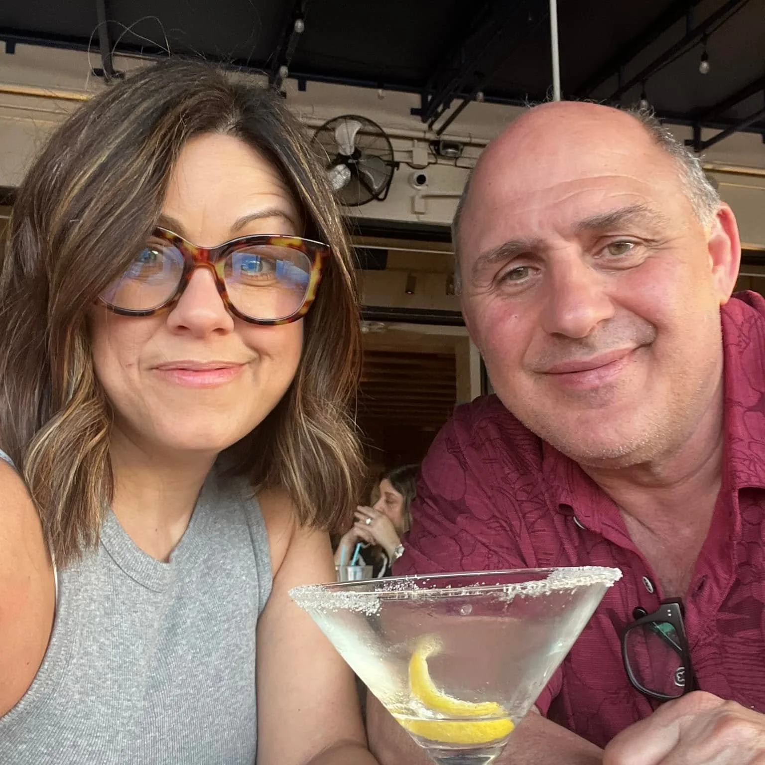 A woman with brown hair and glasses and a man with short hair sitting together at a table, smiling, with a martini glass in front of the woman containing a lemon twist.