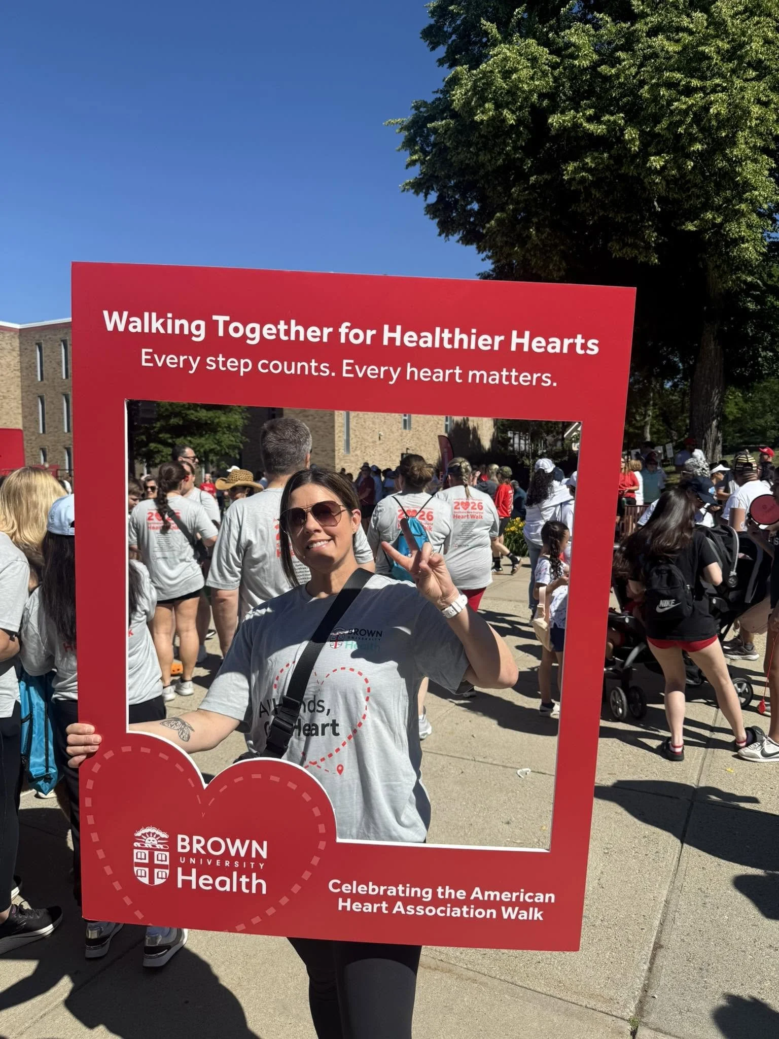 Person holding a red frame with white text that reads, 'Walking Together for Healthier Hearts, Every step counts. Every heart matters,' during a parade or event. The background shows a crowd of people and trees.