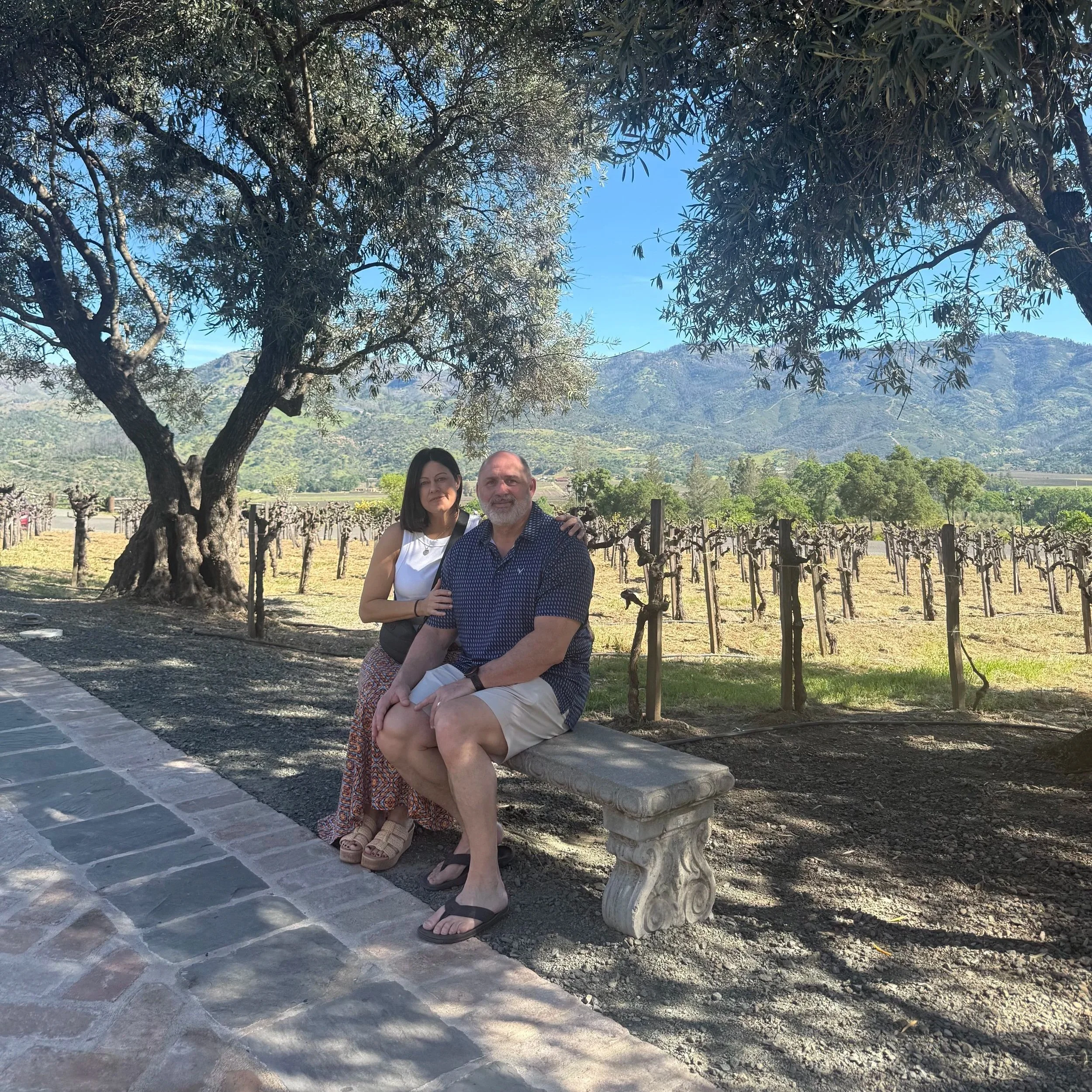 A middle-aged man and woman sit close together on a stone bench under a large tree in a scenic outdoor vineyard or farm setting with mountains in the background on a sunny day.