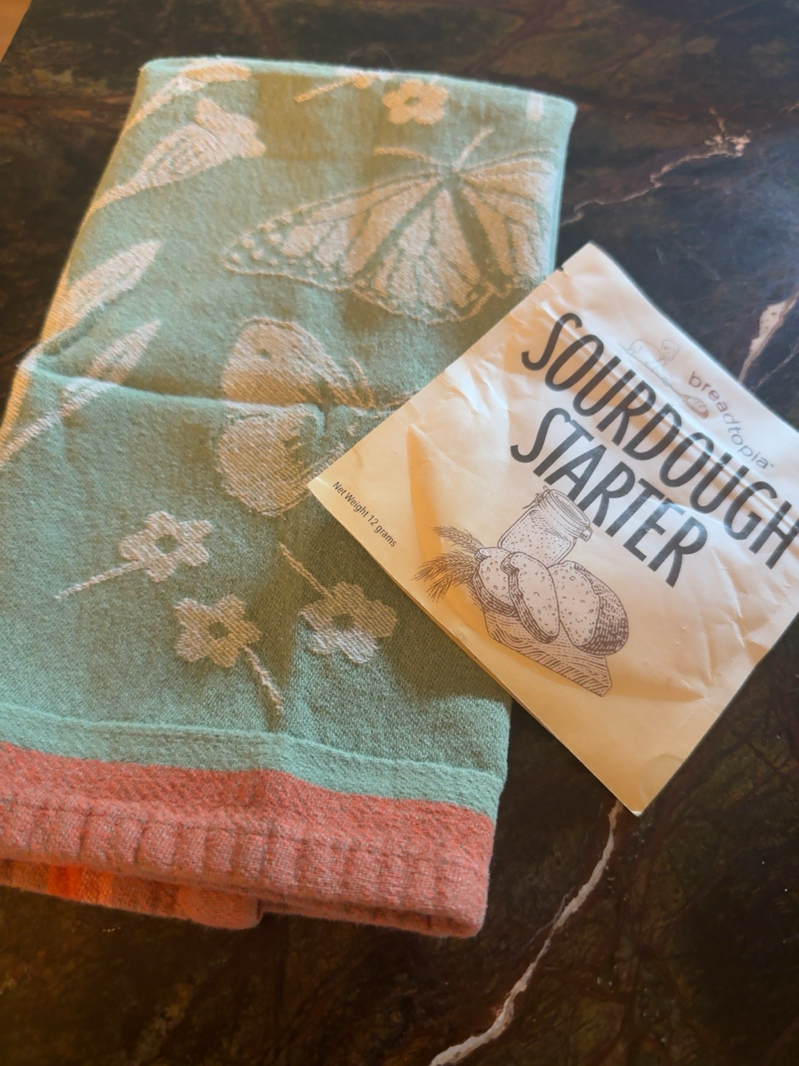 A green dish towel with white floral and butterfly embroidery is next to a packet labeled Sour Dough Starter. The packet has a black and white illustration of bread, a jar, and wheat on it. They are placed on a dark, marbled surface.