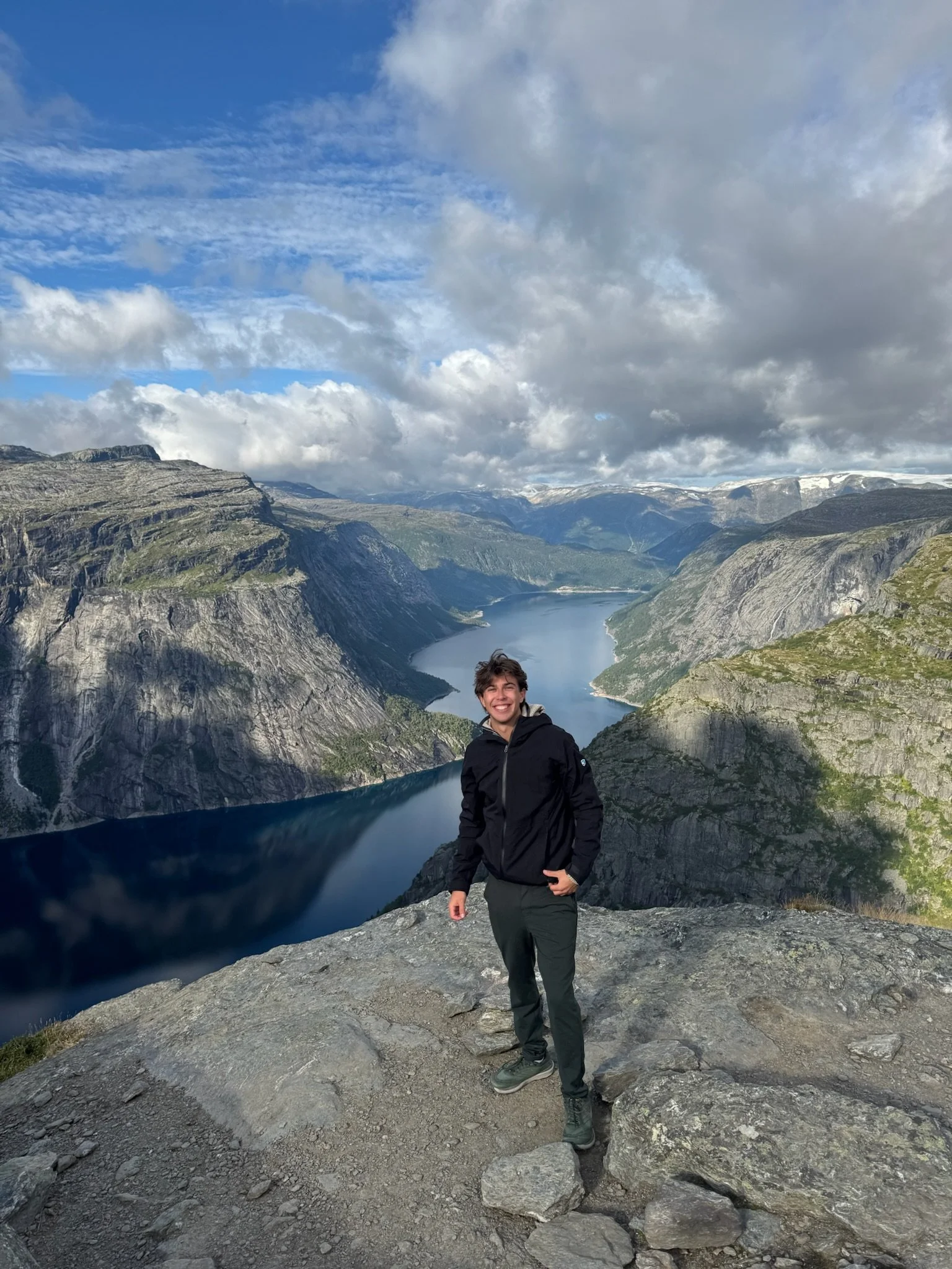 A young man with dark hair smiling and standing on a rocky overlook with a large body of water, mountains, and a partly cloudy sky in the background.