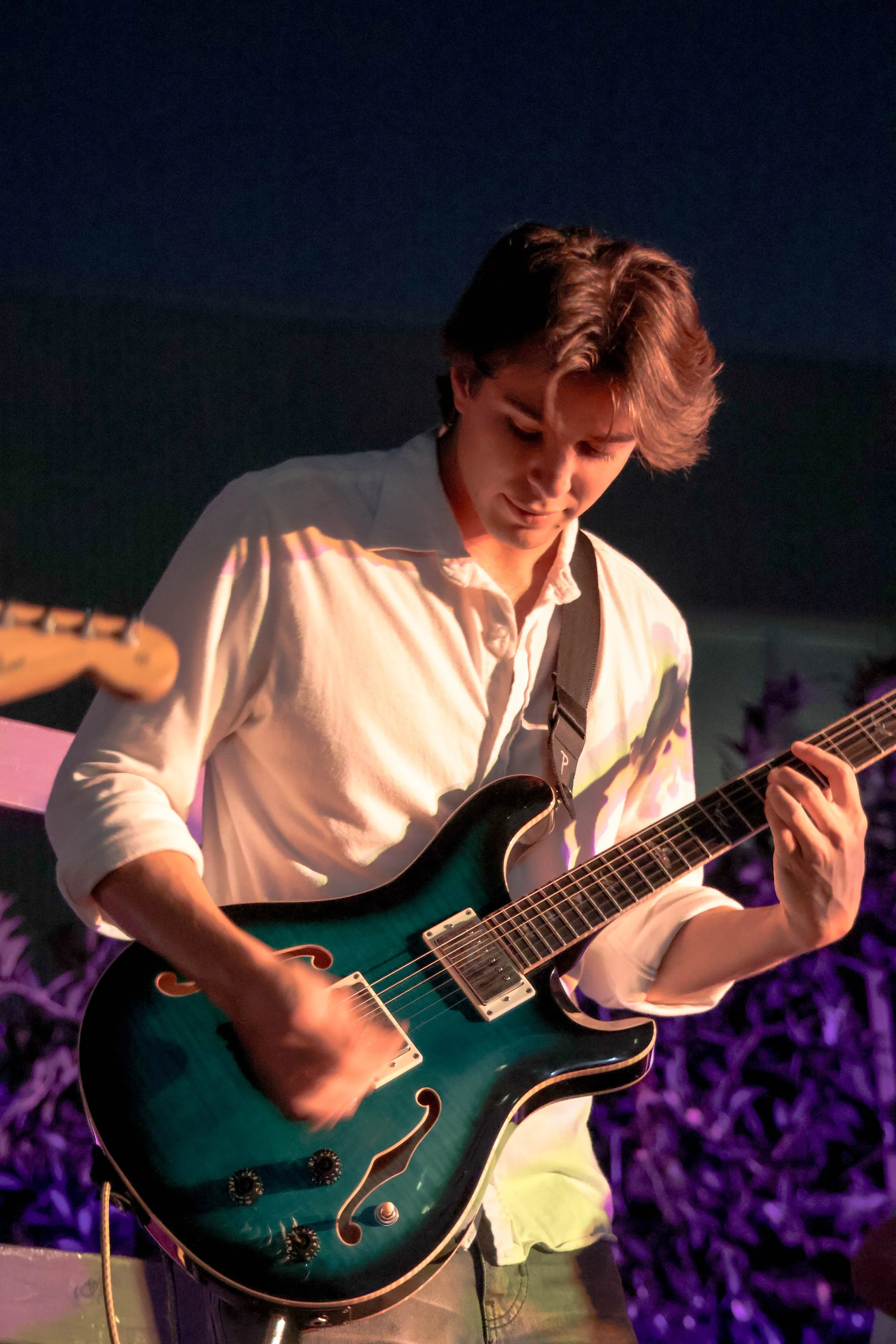 A young man with wavy brown hair playing a teal electric guitar on stage at night, with purple lighting and blurred plants in the background.