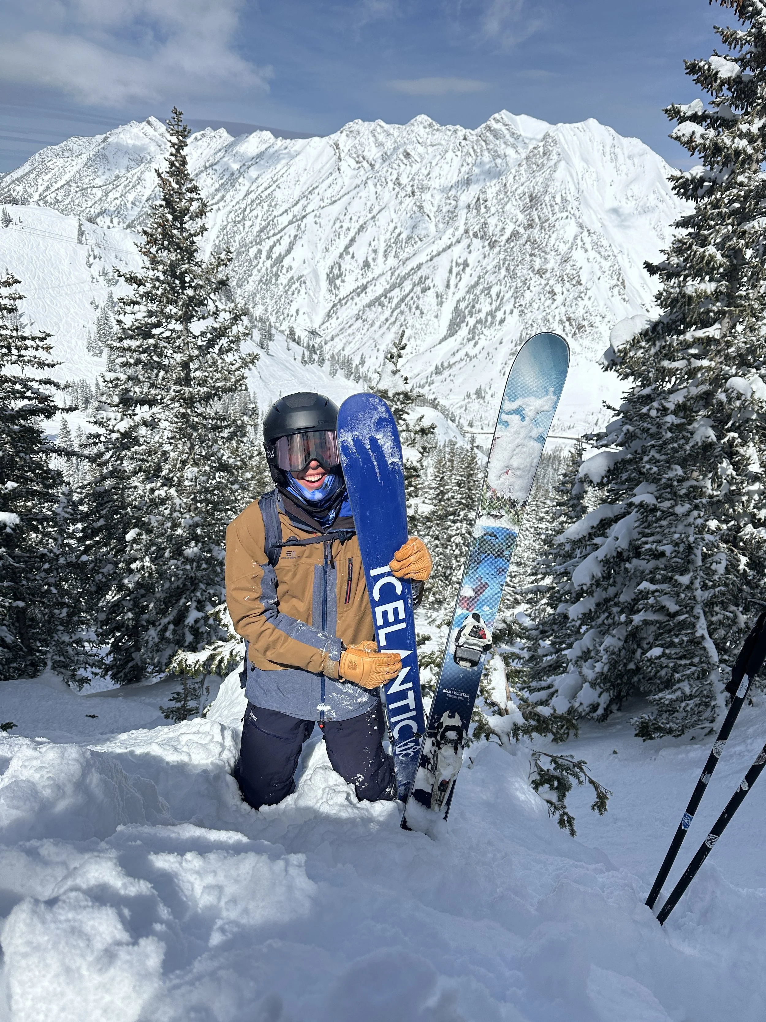 A person dressed in winter gear, including a helmet, goggles, and gloves, holding blue ski equipment and standing in deep snow on a snowy mountain landscape with trees and high mountain peaks in the background.
