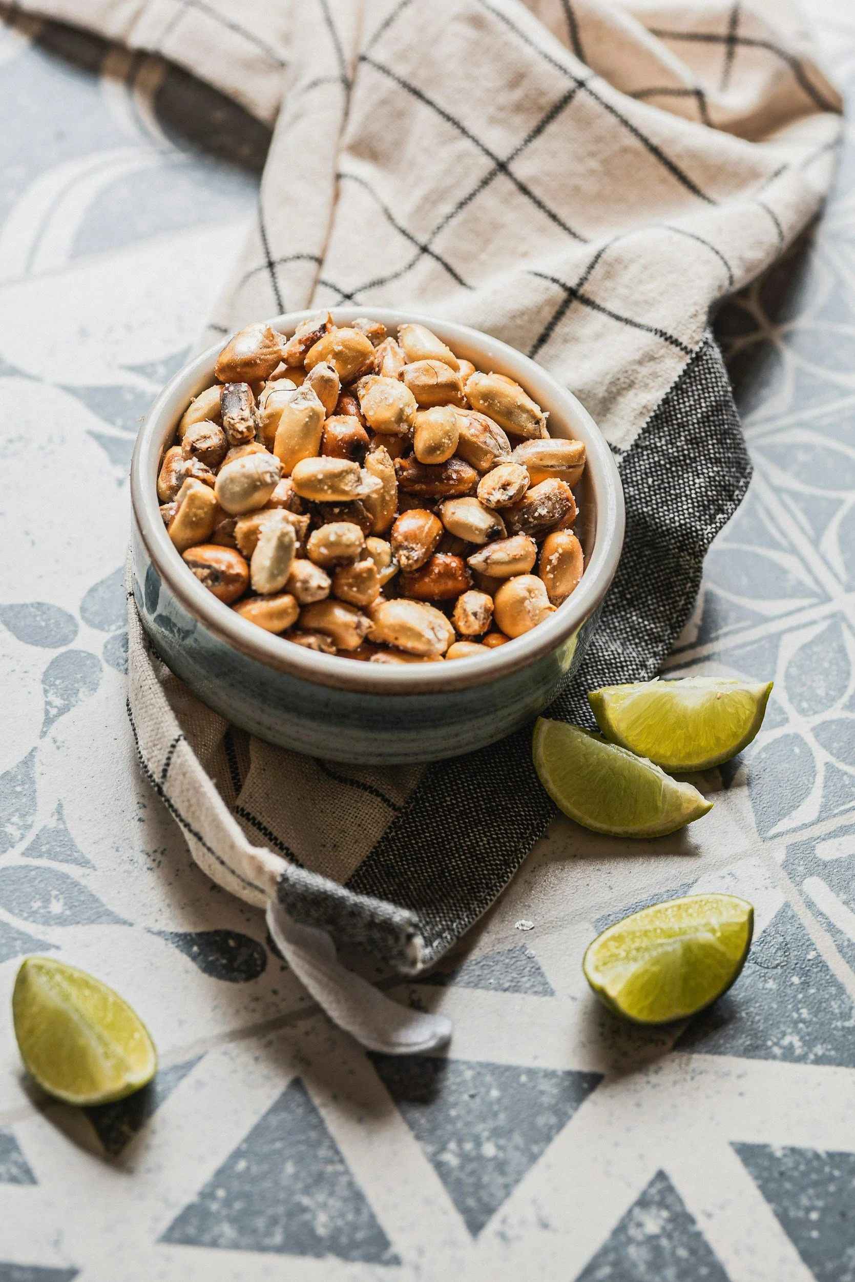 A bowl of salted peanuts on a checkered cloth with lime wedges around it, on a decorative table.