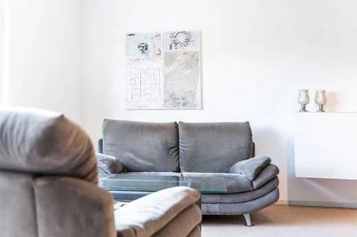 Wohnzimmer mit grauem Sofa, weißen Wänden, moderner Kunst an der Wand und zwei Lampen auf einem weißen Regal.