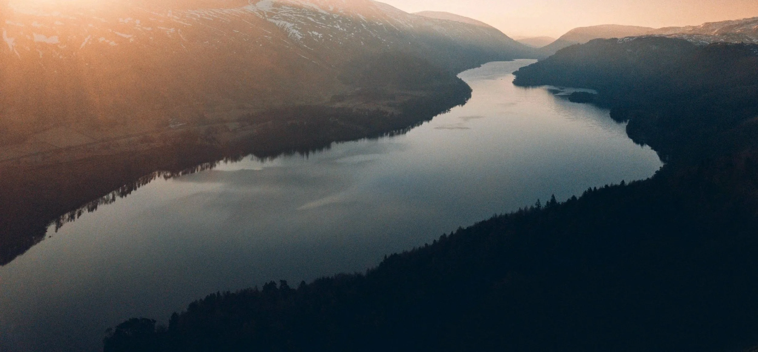 Sunny day over a winding river surrounded by forested mountains with snow patches.