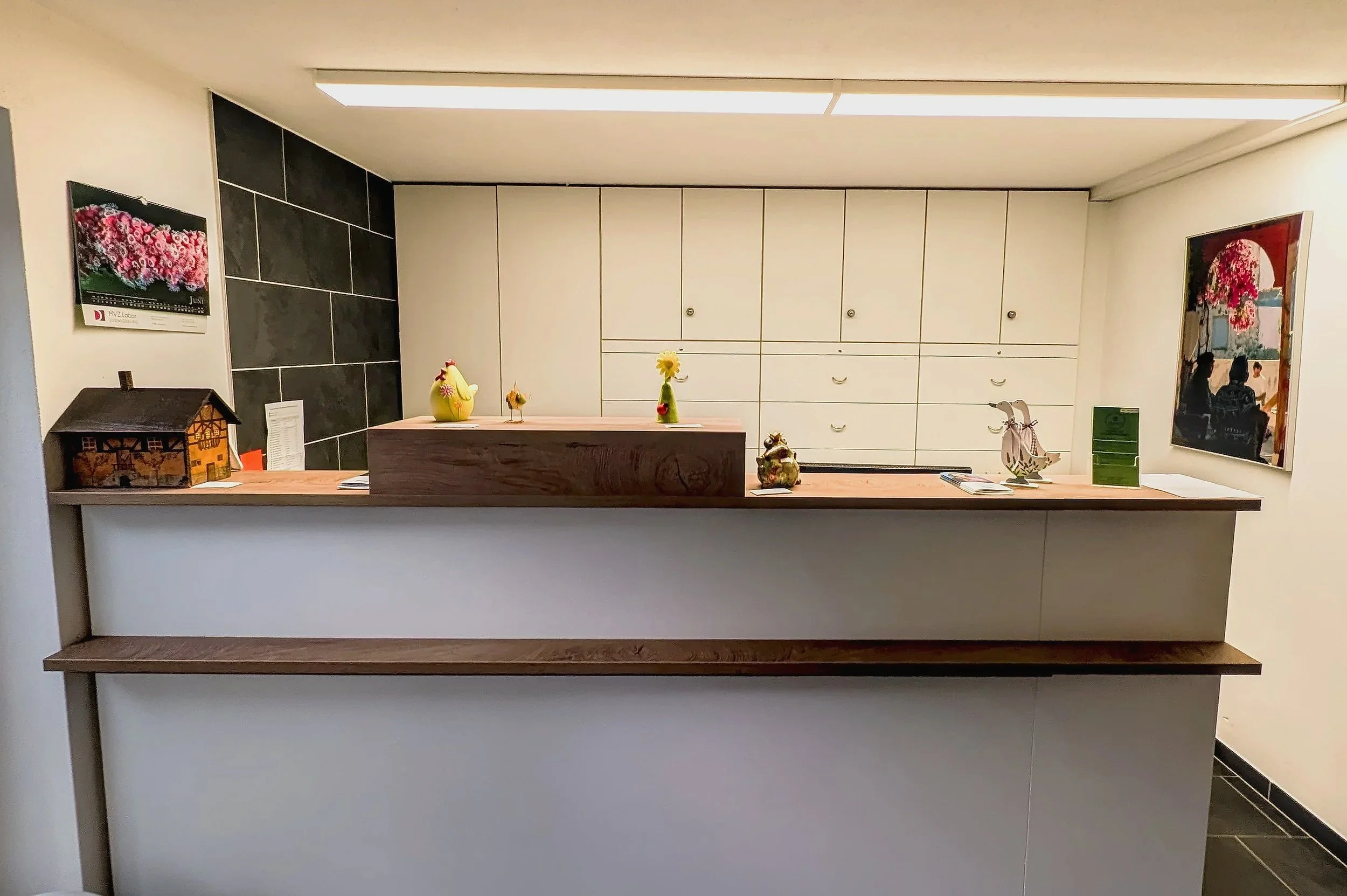 Ein Empfangstresen mit Dekorationsartikeln und weißen Schränken im Hintergrund. An der Wand hängen zwei Fotos, eines mit Blumen und eines mit Personen auf einer Bank im Freien.