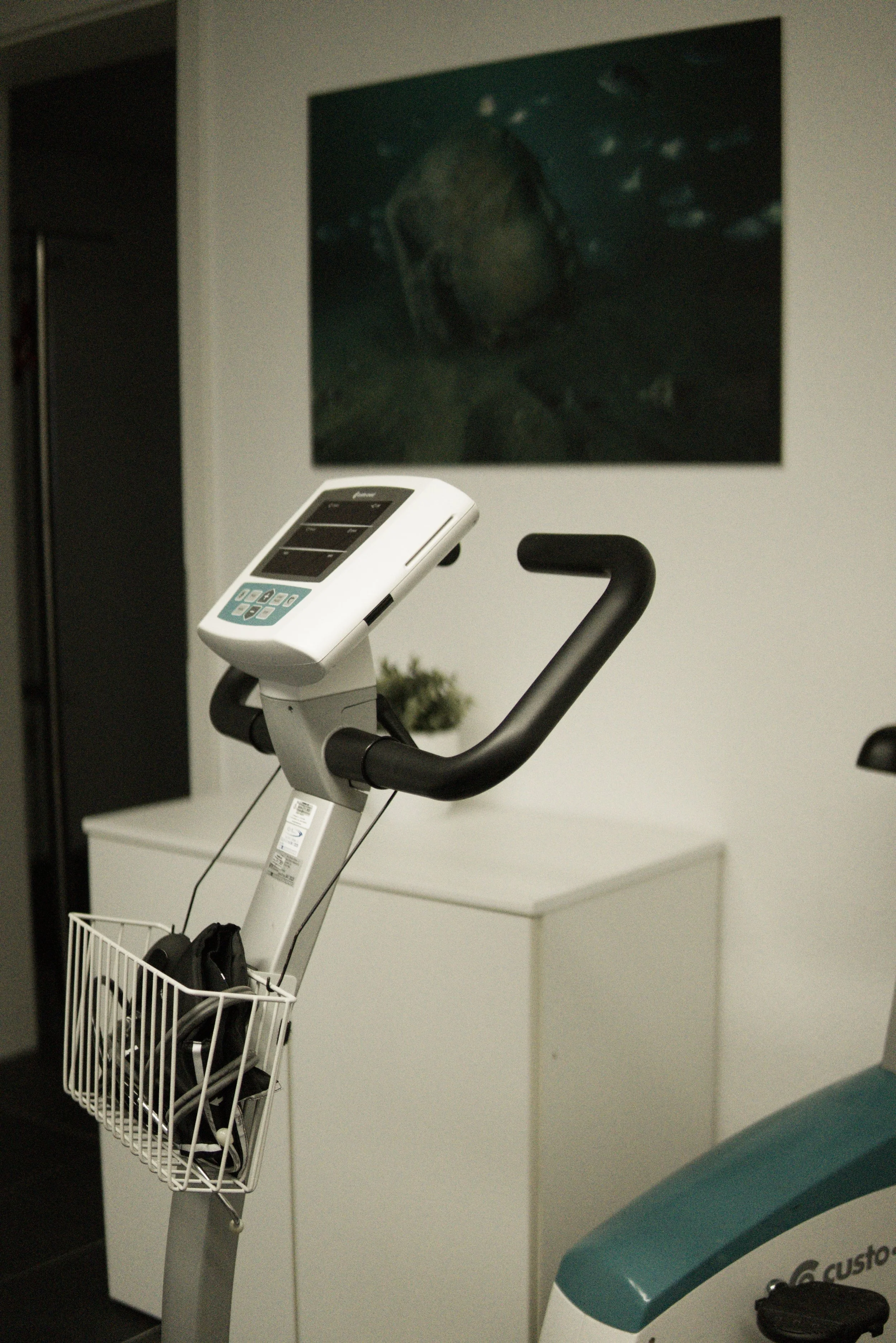 Ein stationäres Fitnessgerät mit einem digitalen Display und einem Fahrradlenker, neben einem weißen Schreibtisch und einem Bild an der Wand.