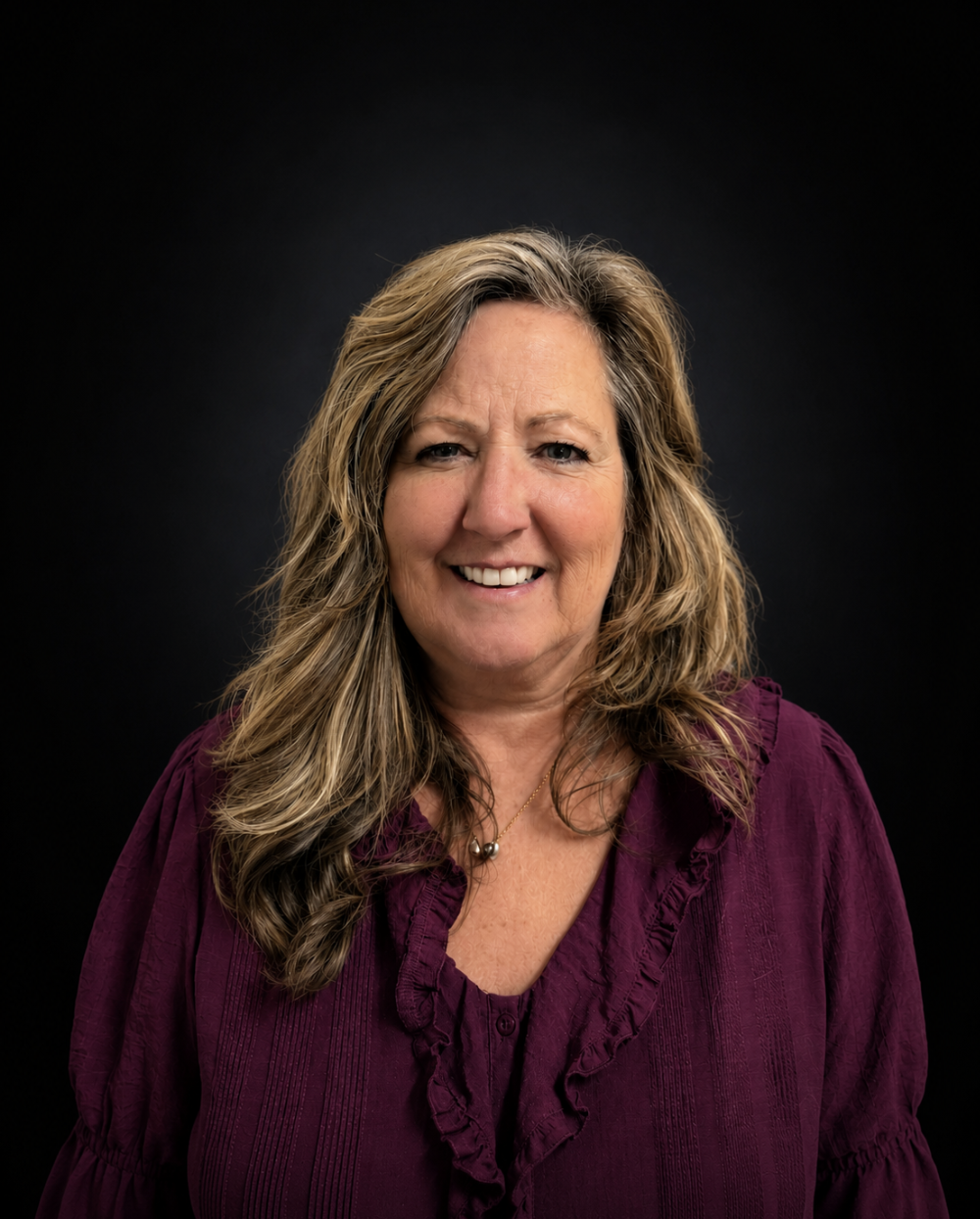 Portrait of a smiling middle-aged woman with long, wavy blonde hair, wearing a purple blouse with ruffles and a pearl necklace, against a black background.