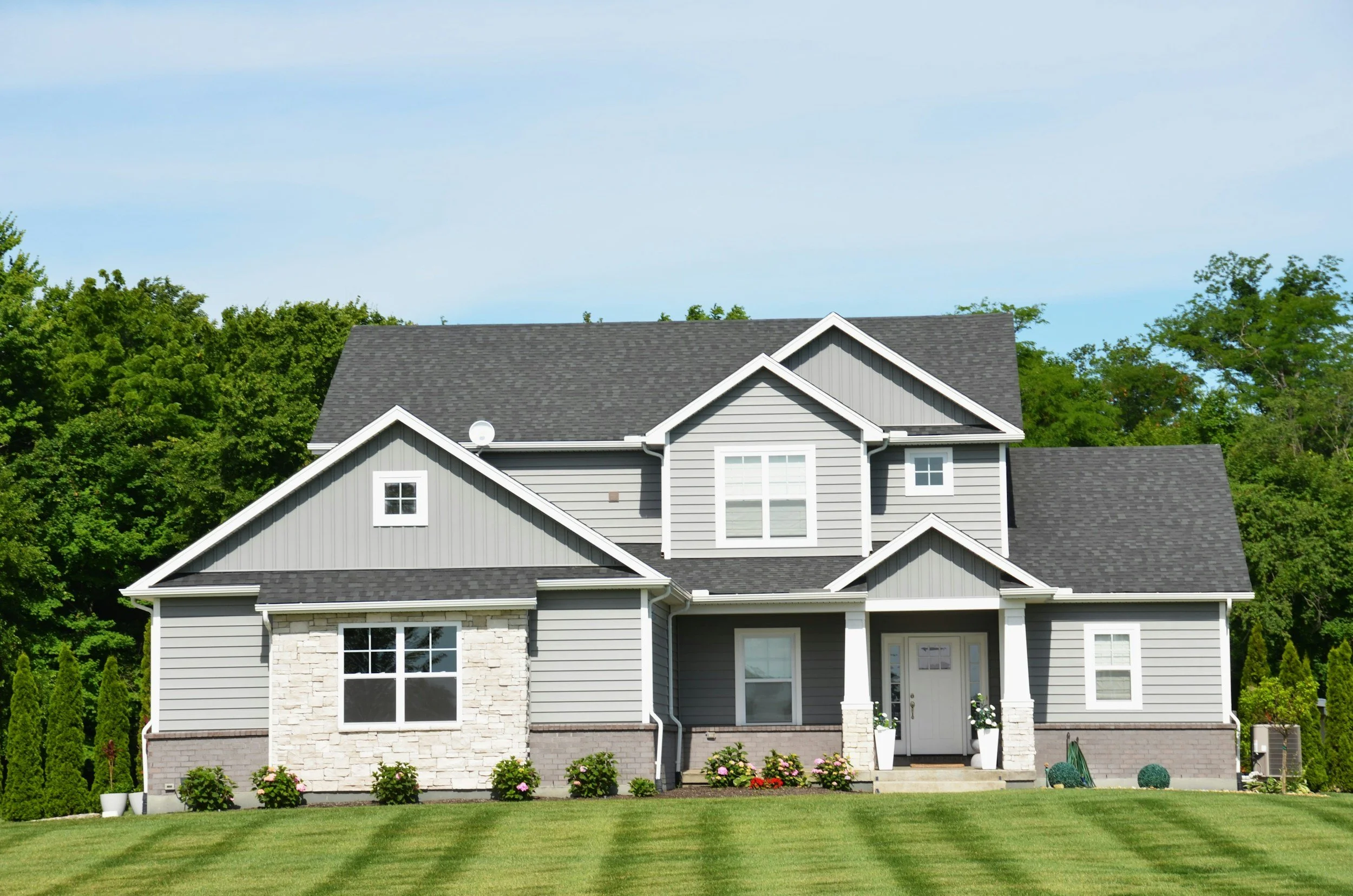 A modern two-story house with gray siding, white trim, and a gray shingled roof, surrounded by a well-maintained lawn and greenery.