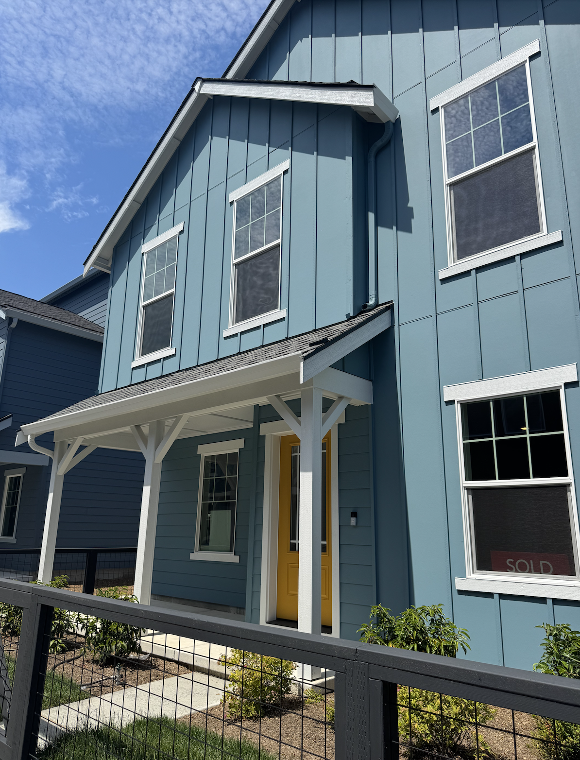 Blue two-story house with white trim and yellow front door, small front porch with white supports, fenced yard, and a 'sold' sign in the window. Clear sky with some clouds.