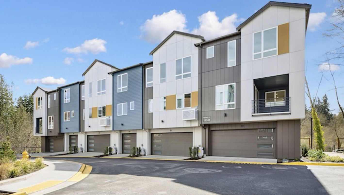 Modern multi-family homes with garage doors on the ground level, painted in various shades of blue, gray, and white, with some windows having balconies, under a partly cloudy sky.